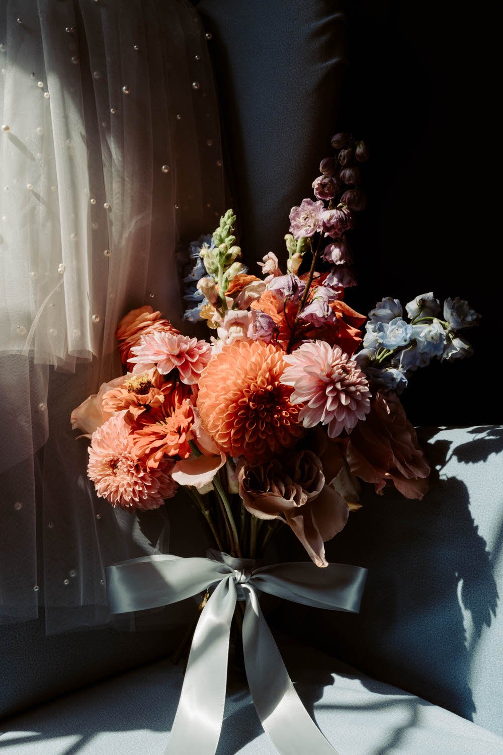 A bouquet of pink, peach, and purple flowers tied with a silver ribbon, placed on a dark surface with a sheer curtain with pearl embellishments in the background.