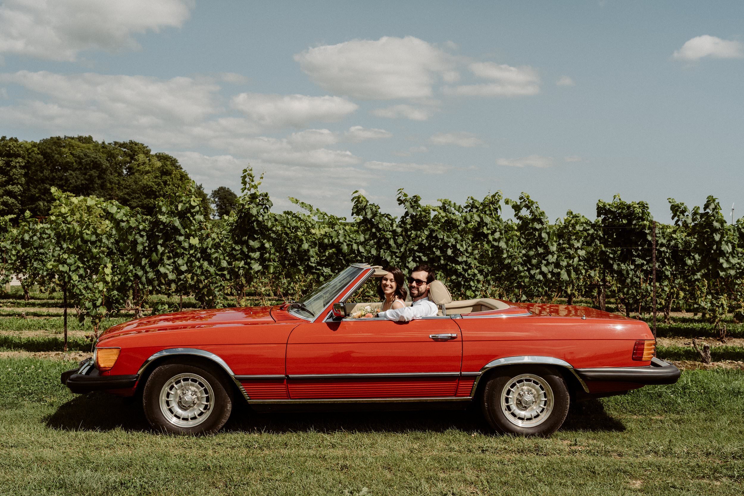A smiling couple in wedding attire driving a red convertible car through a vineyard under a partly cloudy sky.