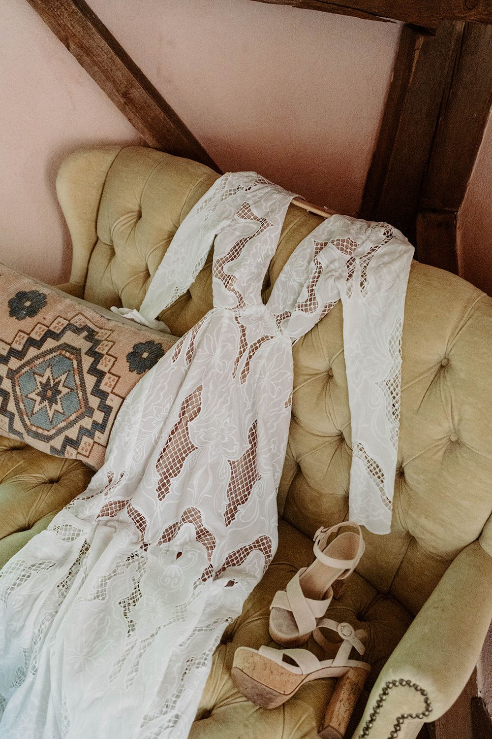 A beige tufted sofa with white lace fabric draped over the backrest, a patterned textile with geometric and star designs on the seat, and a pair of nude high-heeled sandals with ankle straps resting on the sofa.