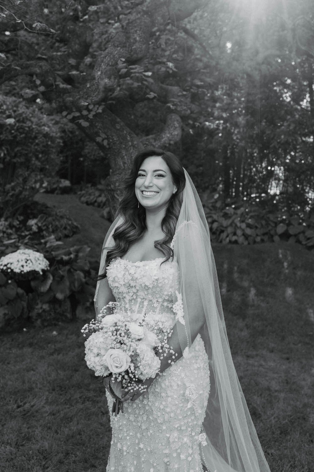 A smiling bride in a lace wedding dress holding a bouquet of flowers outdoors near a large tree.