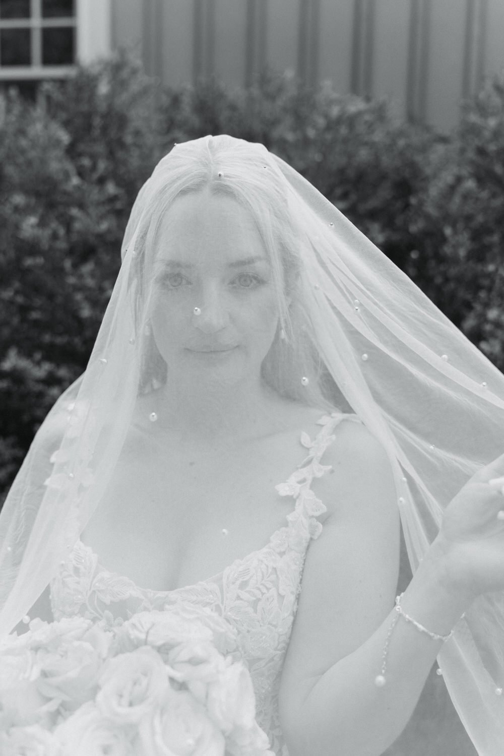A bride in a wedding dress with lace details, wearing a veil, holding a bouquet of flowers, standing outdoors with greenery in the background.
