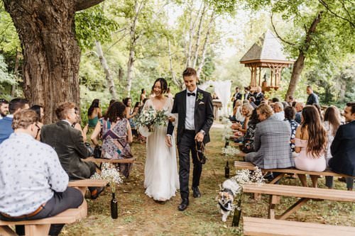 Bride and groom walking down the aisle at outdoor wedding ceremony in a wooded area, surrounded by seated guests, with a gazebo in the background.