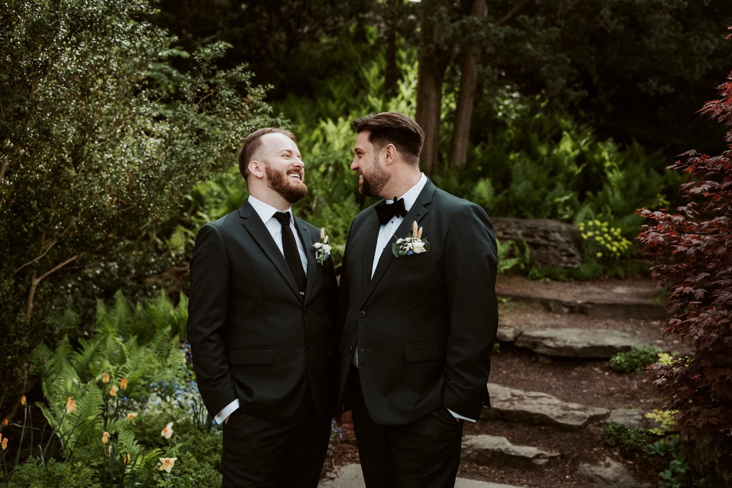 Two men in tuxedos, holding hands and smiling at each other, standing in a lush green garden.