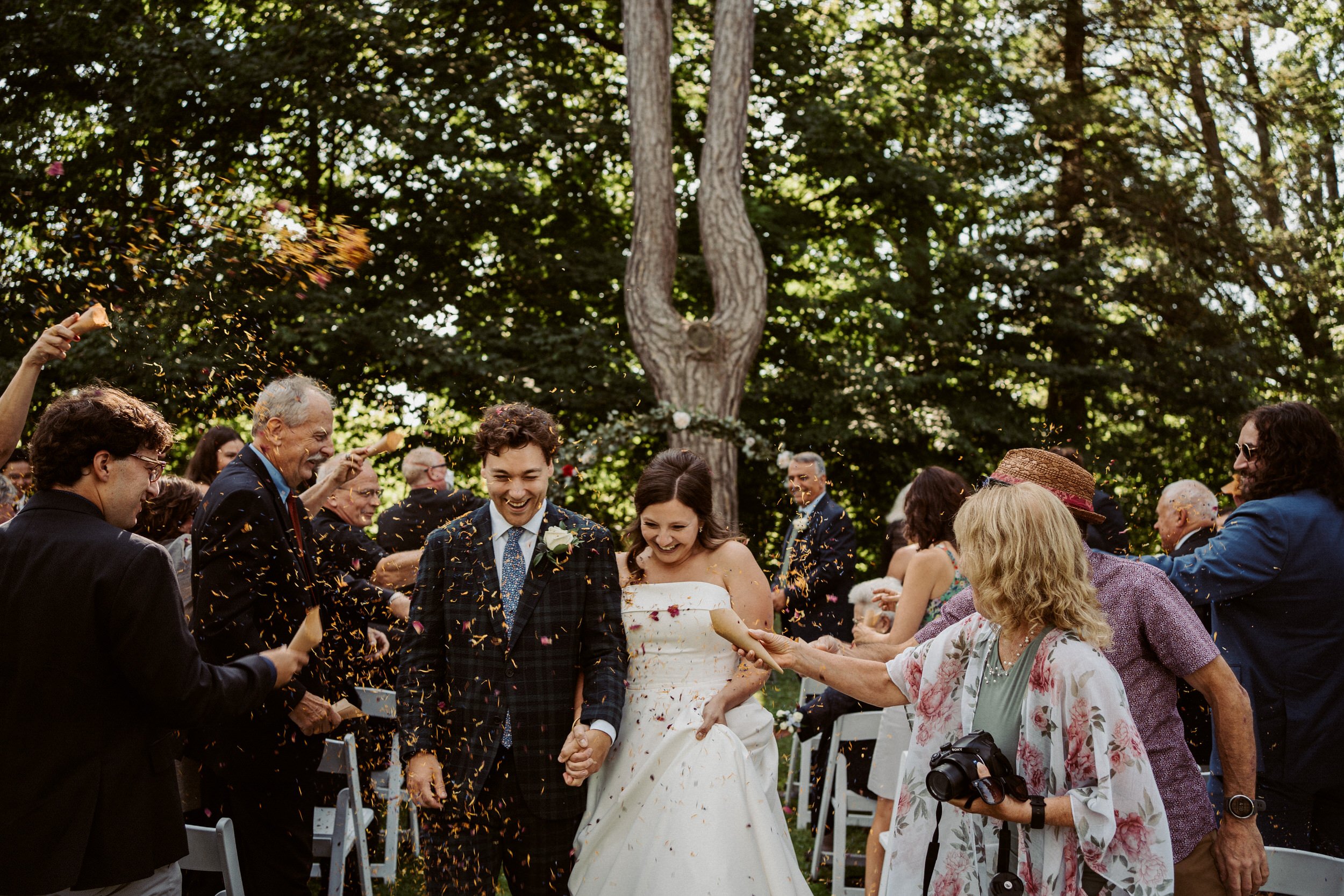 A group of people celebrating a wedding outdoors, some are throwing confetti as the bride and groom smile and hold hands, surrounded by trees.