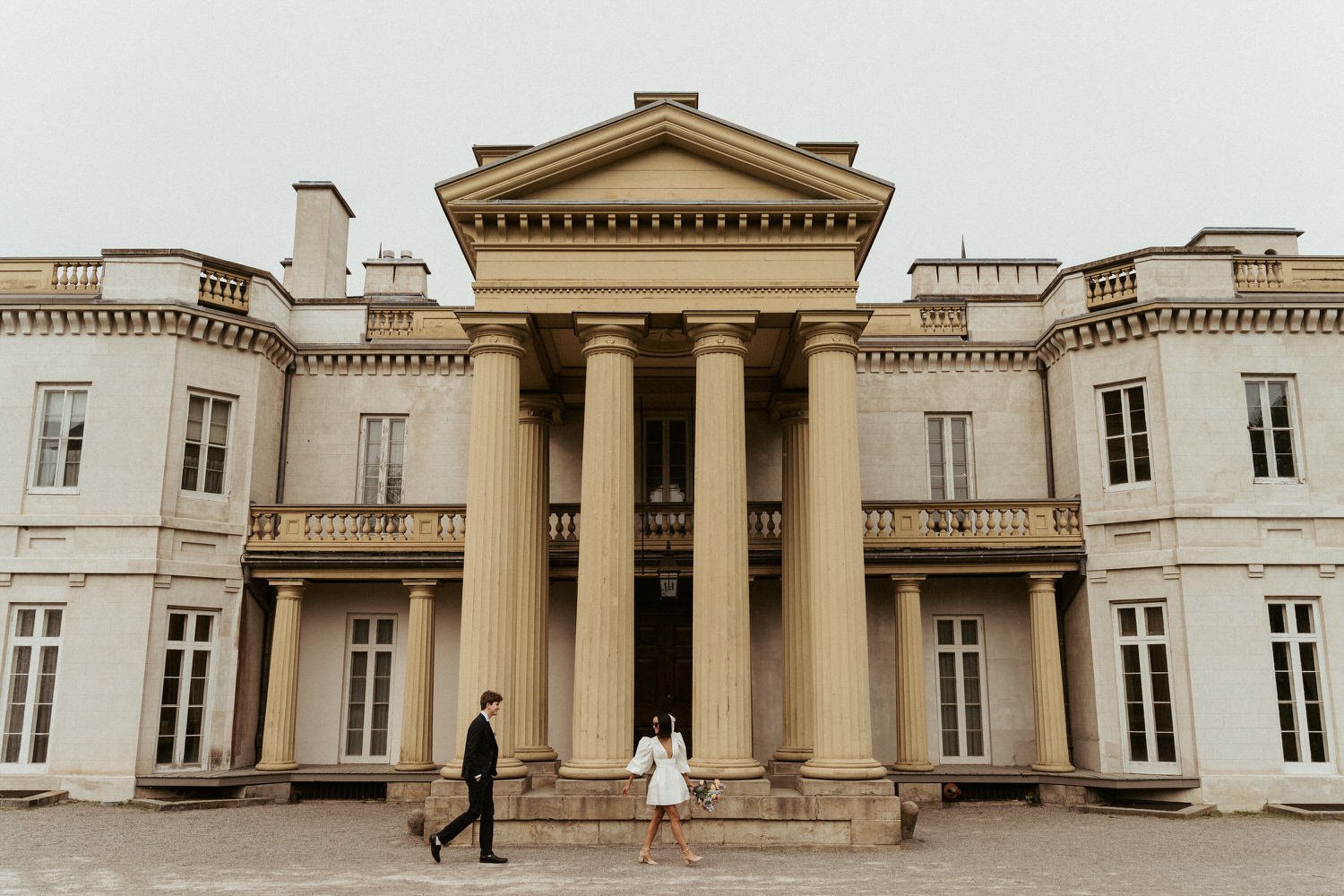 A wedding couple standing in front of a large, historic mansion with classical columns and ornate architectural details.