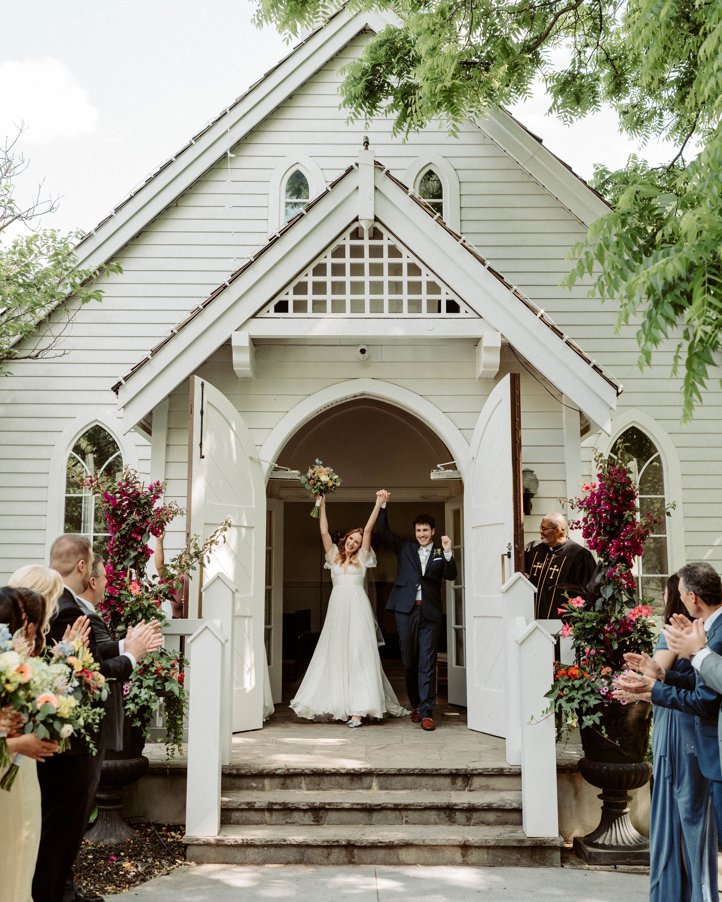 A newlywed couple holding hands and celebrating as they exit a white chapel, surrounded by guests, during a wedding ceremony. The bride holds a bouquet of flowers, and the groom is smiling. Guests are applauding and standing on either side of the entrance, which is decorated with pink and purple flowers, with the open chapel doors behind them.