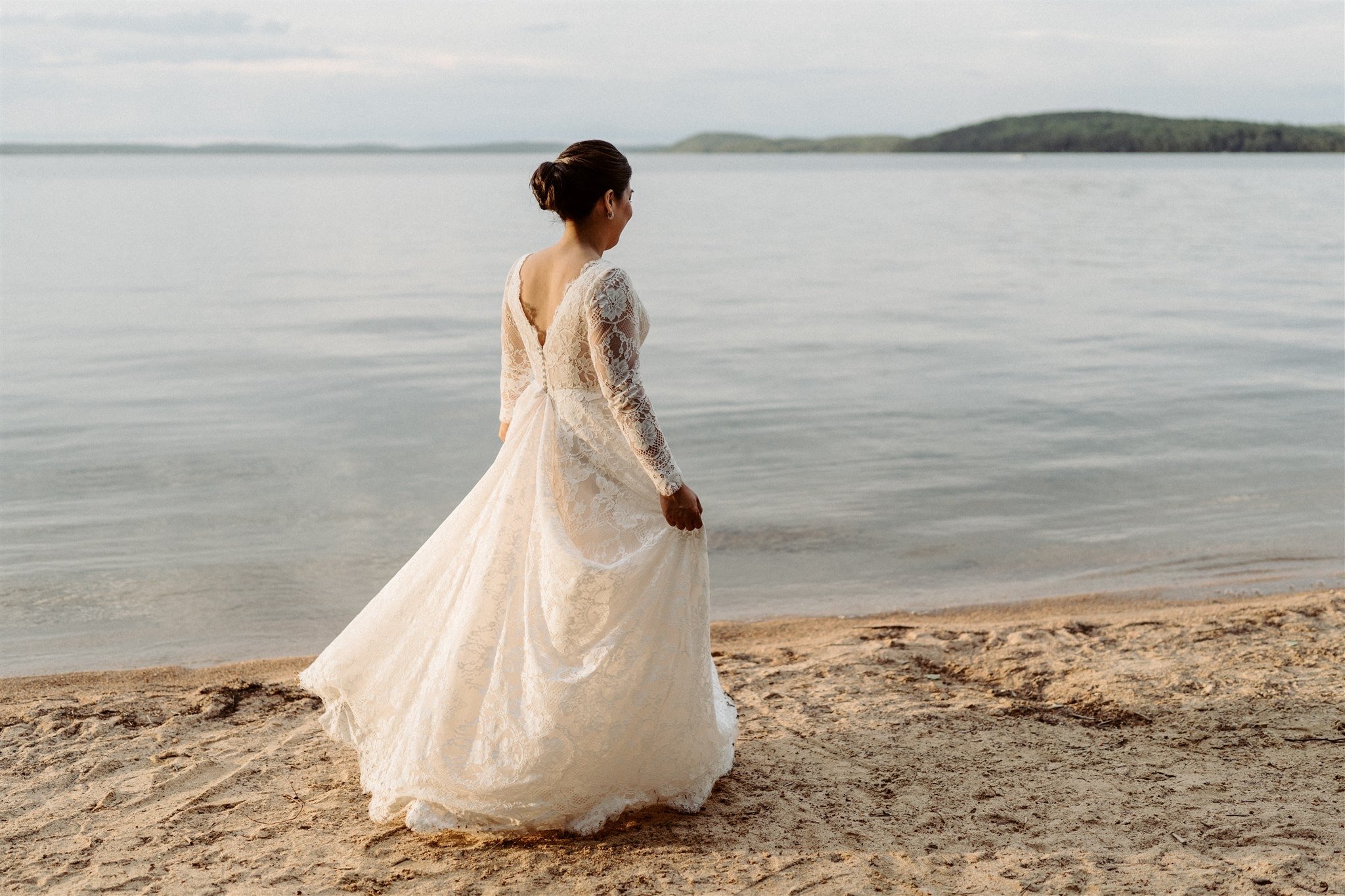 A woman in a white lace wedding dress stands on a sandy beach, looking at the water and distant hills.