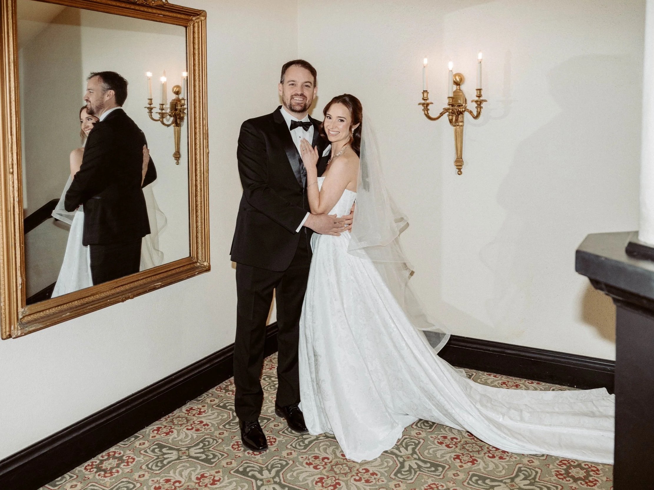 A man and woman dressed in formal wedding attire, standing close together and smiling in a room with a wall mirror and wall-mounted candle holders.