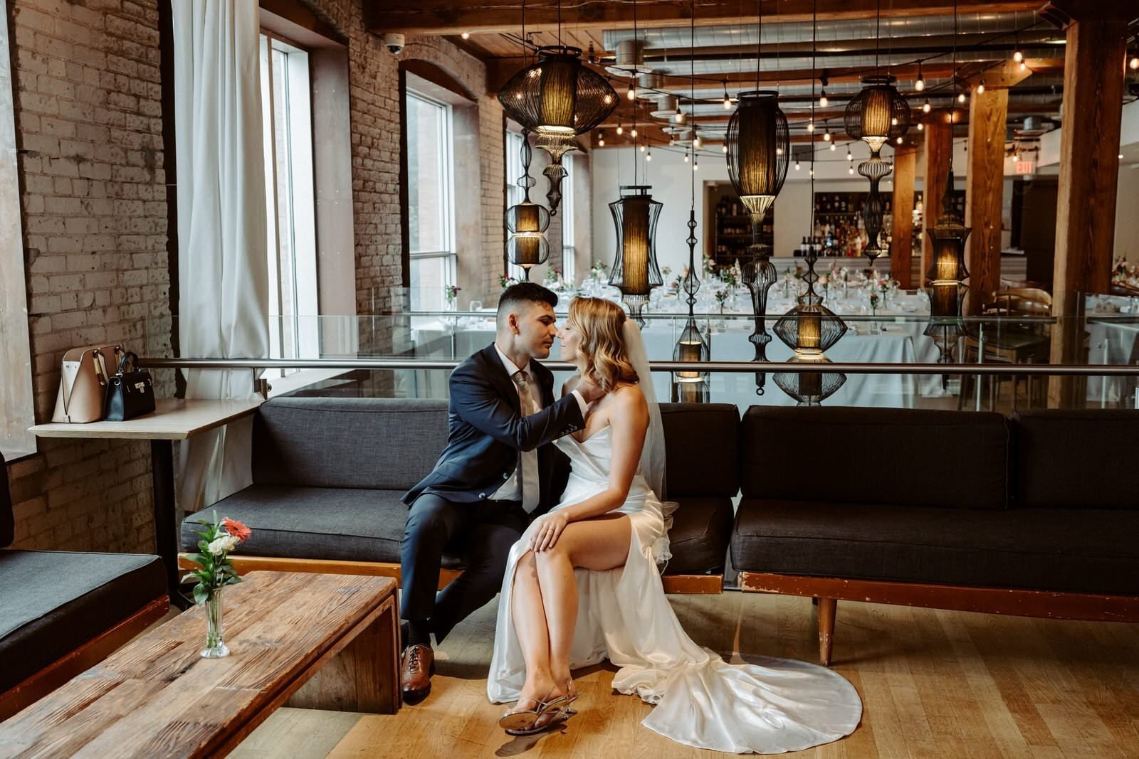 A man and a woman in wedding attire sitting together on a black couch in a rustic indoor setting. The man, dressed in a black suit, gently holds the woman's face while they look into each other's eyes. The woman is wearing a white wedding dress with 