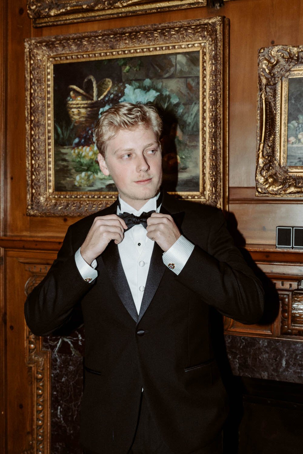 Young man in a tuxedo adjusting his bowtie in front of framed artwork, wooden walls, and a marble mantle.