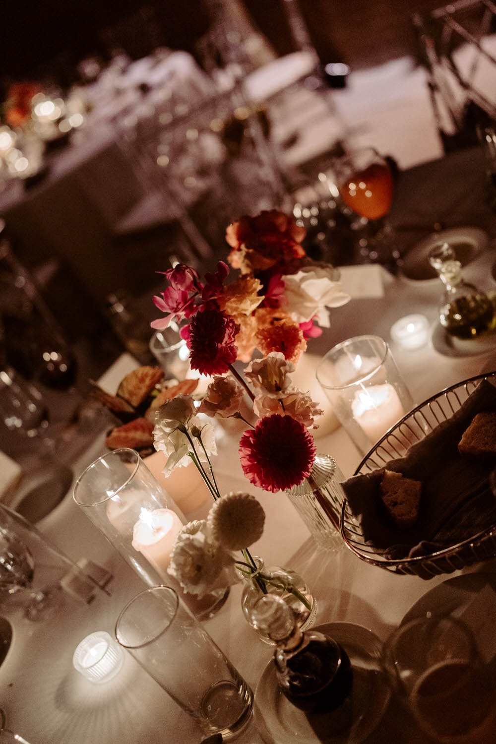 A close-up of a table centerpiece with various flowers in a clear glass vase, surrounded by candlelit glasses and dishes, set in a dimly lit setting ideal for a formal event or dinner party.
