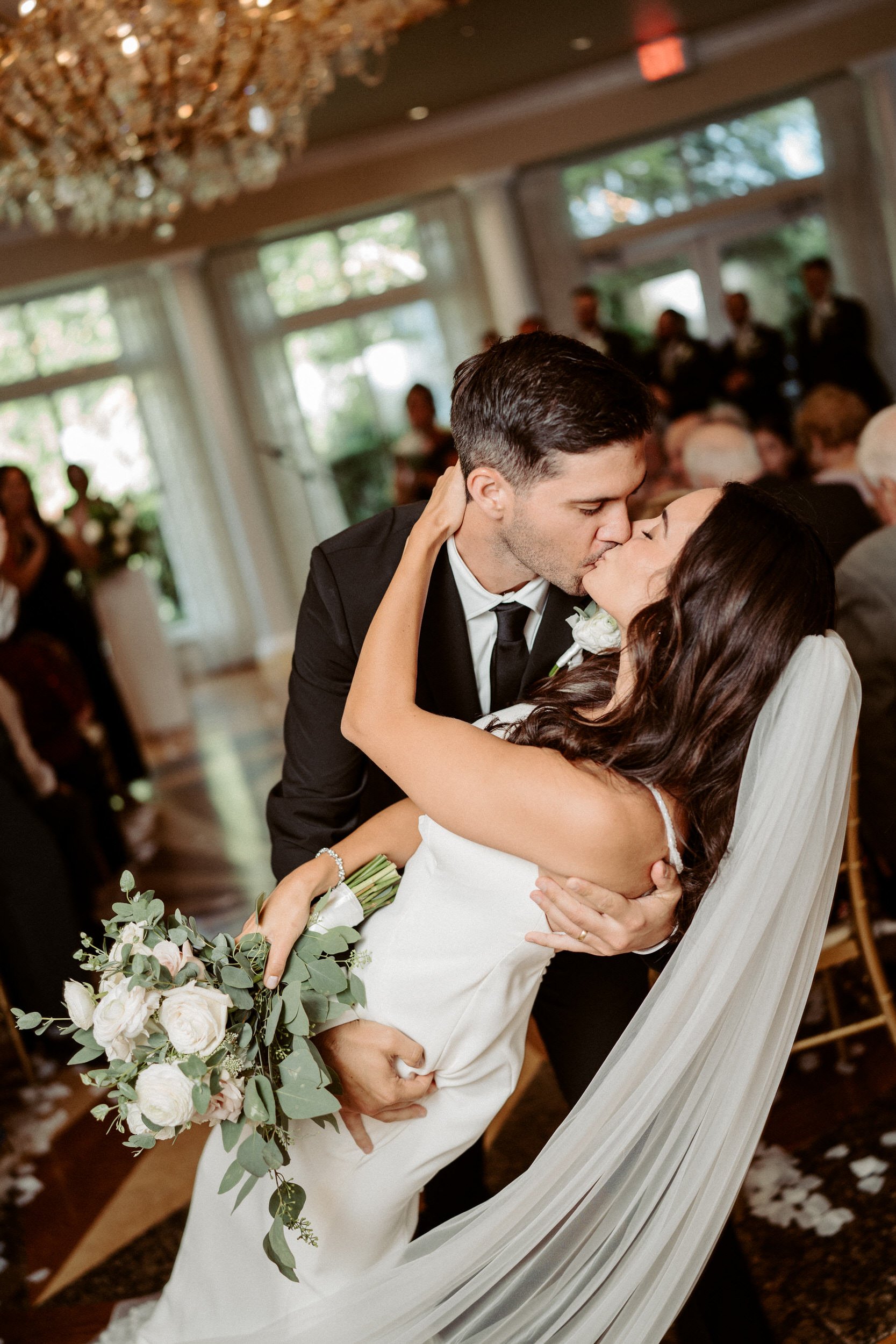 A newlywed couple shares a kiss during their wedding reception, with guests and a chandelier visible in the background.