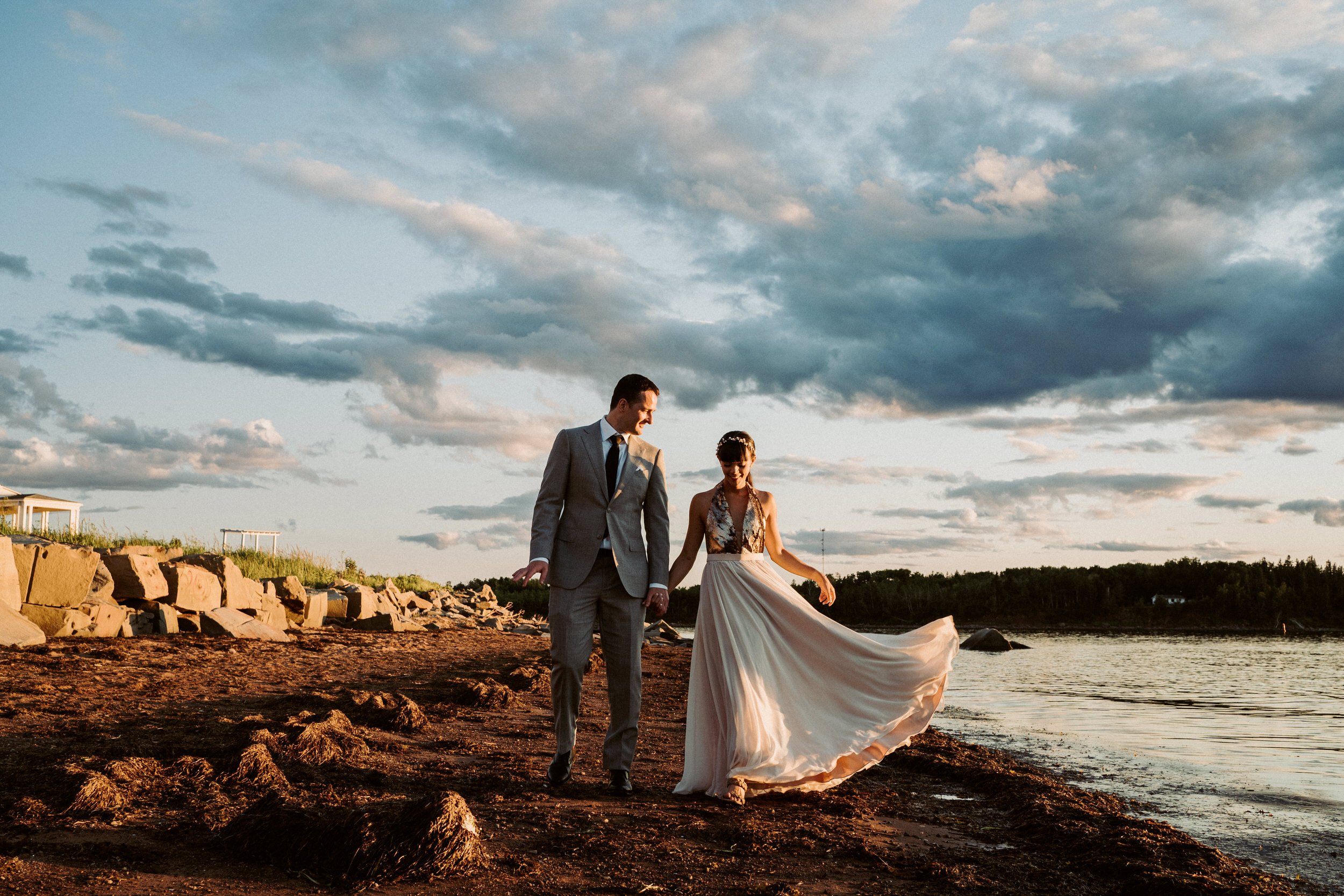 A couple dressed in wedding attire walking along a rocky shoreline at sunset, holding hands and smiling, with a partly cloudy sky overhead.