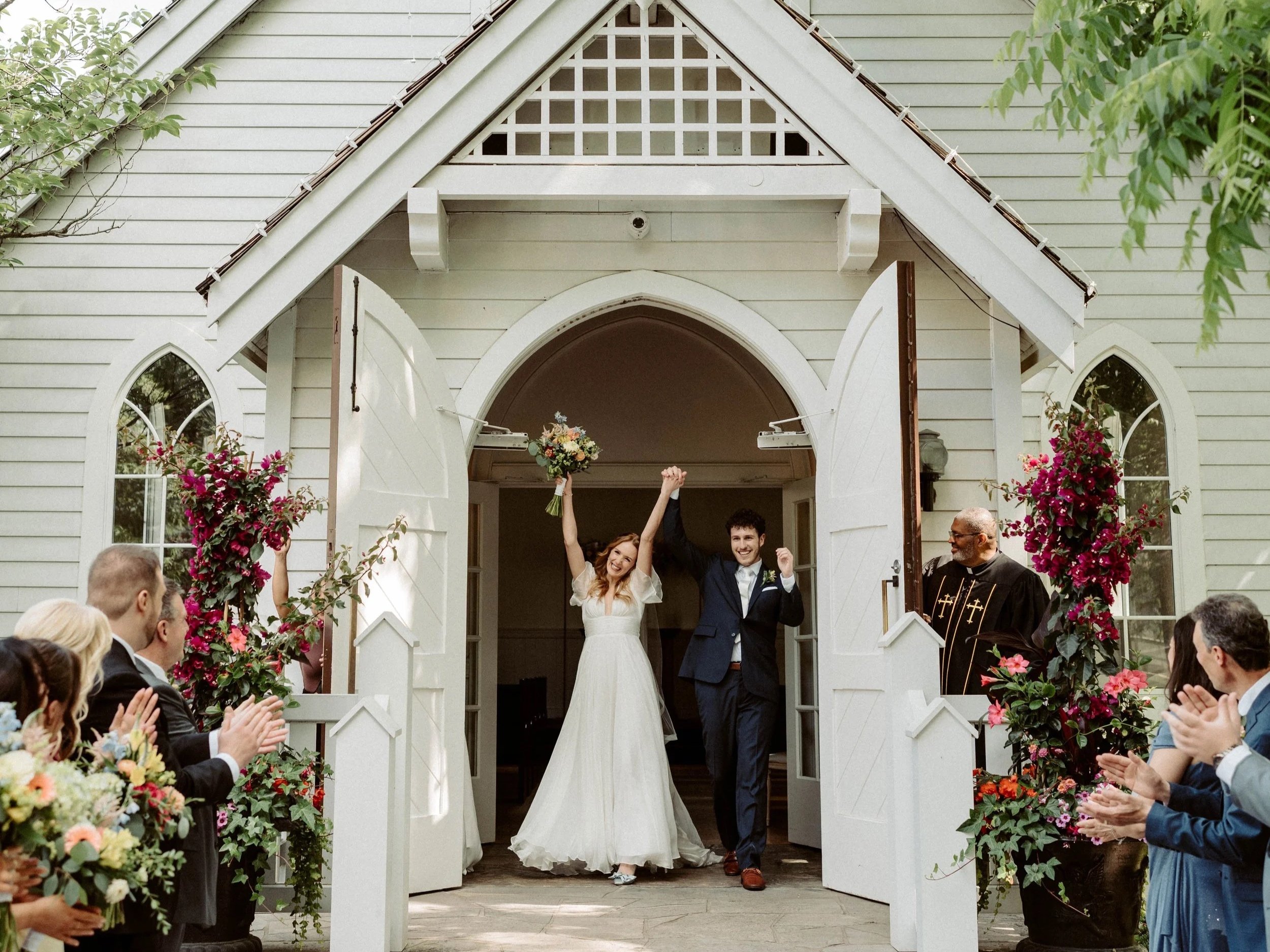 A wedding ceremony taking place outside a white church with an arched entrance. The bride and groom are holding hands inside the doorway, smiling and celebrating as guests sit and applaud outside. The bride is holding a bouquet, and the groom is dressed in a dark suit. The scene is decorated with flowers and greenery.