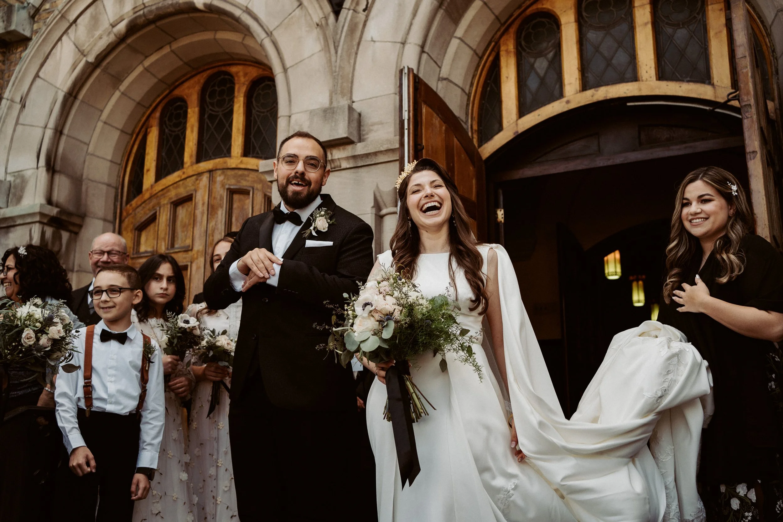 A wedding scene with a bride and groom smiling and holding hands, surrounded by friends and family, outside of a church with large wooden doors and stone architecture.