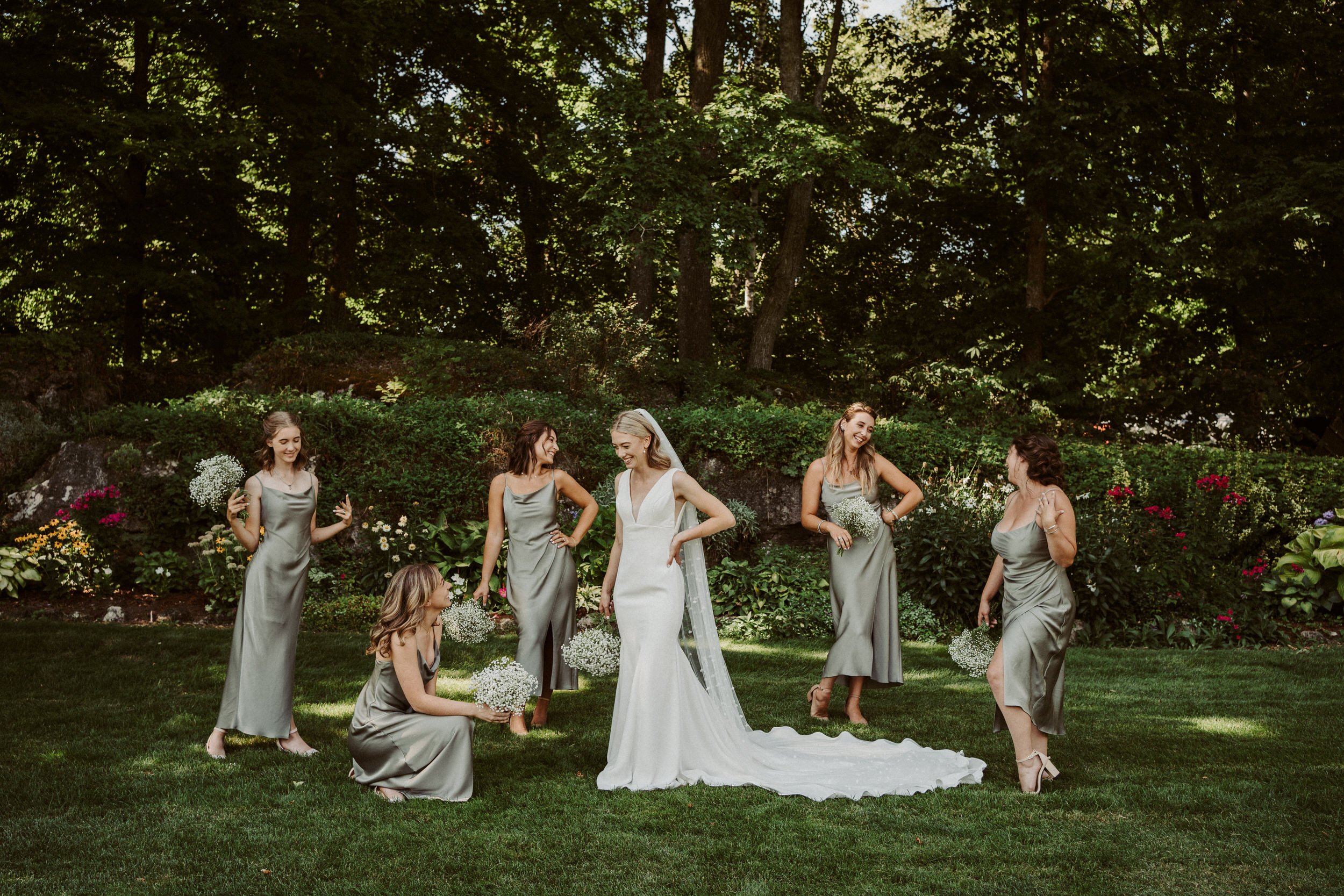 A bride and her bridesmaids standing on a lush green lawn during a wedding celebration, surrounded by trees and colorful flowers.