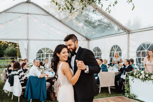 Bride and groom dancing at wedding reception under a clear tent with guests seated at tables and twinkle lights overhead.