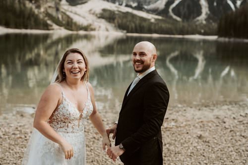 A bride and groom holding hands and smiling at the camera on a rocky shoreline with a calm lake and snow-capped mountains in the background.