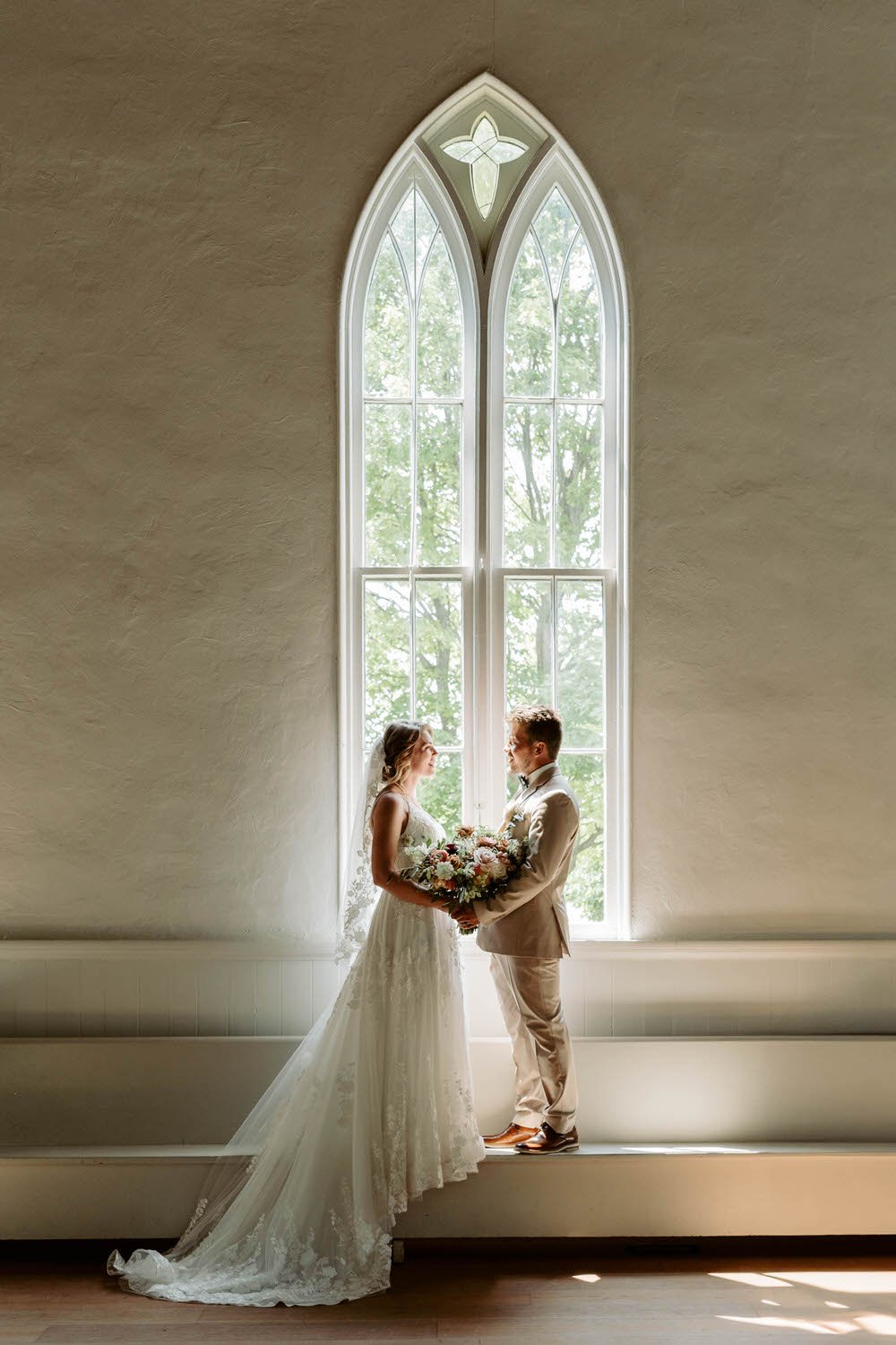 Bride and groom standing face to face in front of a tall window in a church, holding a bouquet of flowers, during their wedding ceremony.