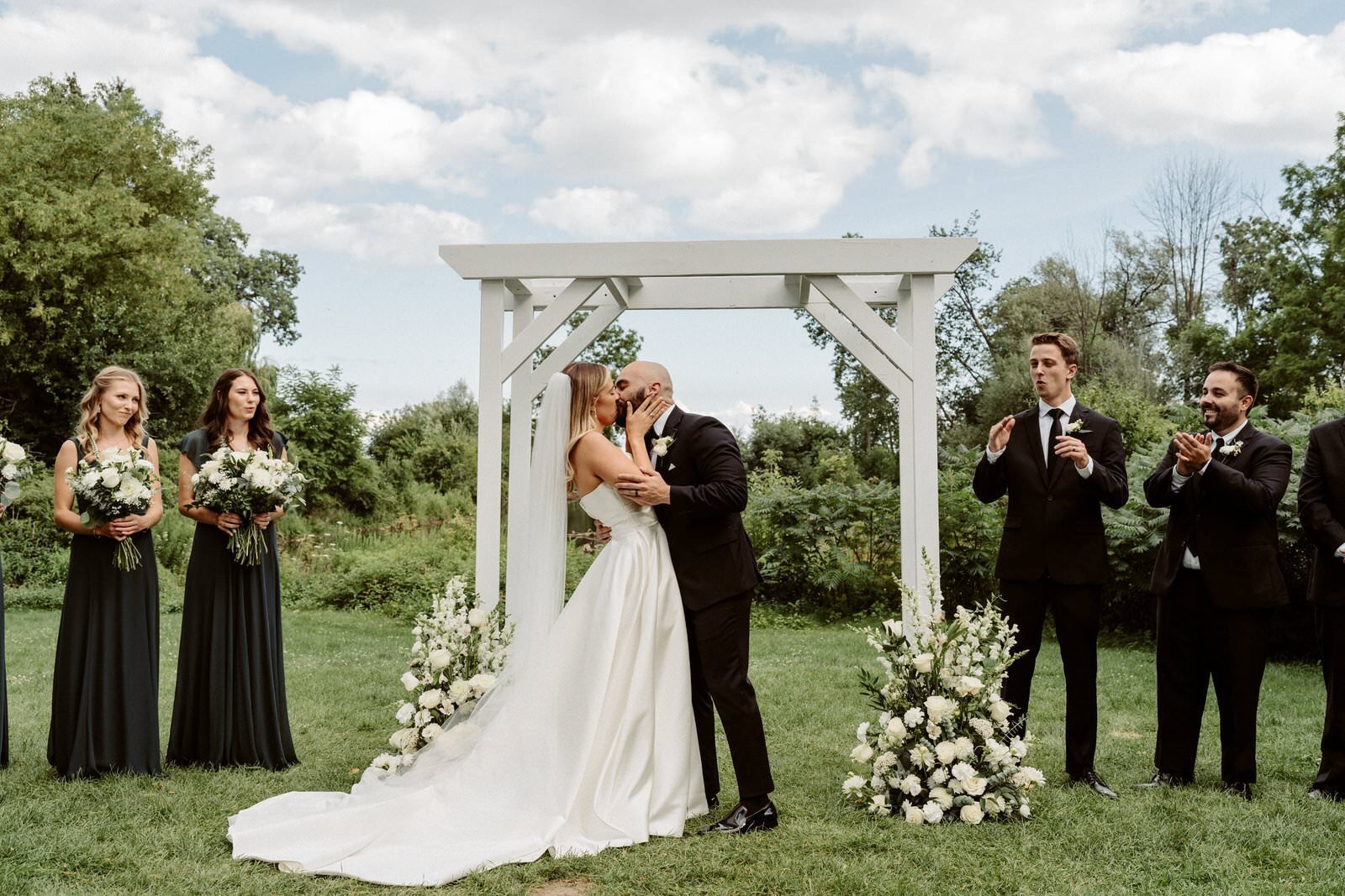 A bride and groom kissing under a white wooden wedding arch surrounded by wedding party and floral arrangements outdoors.