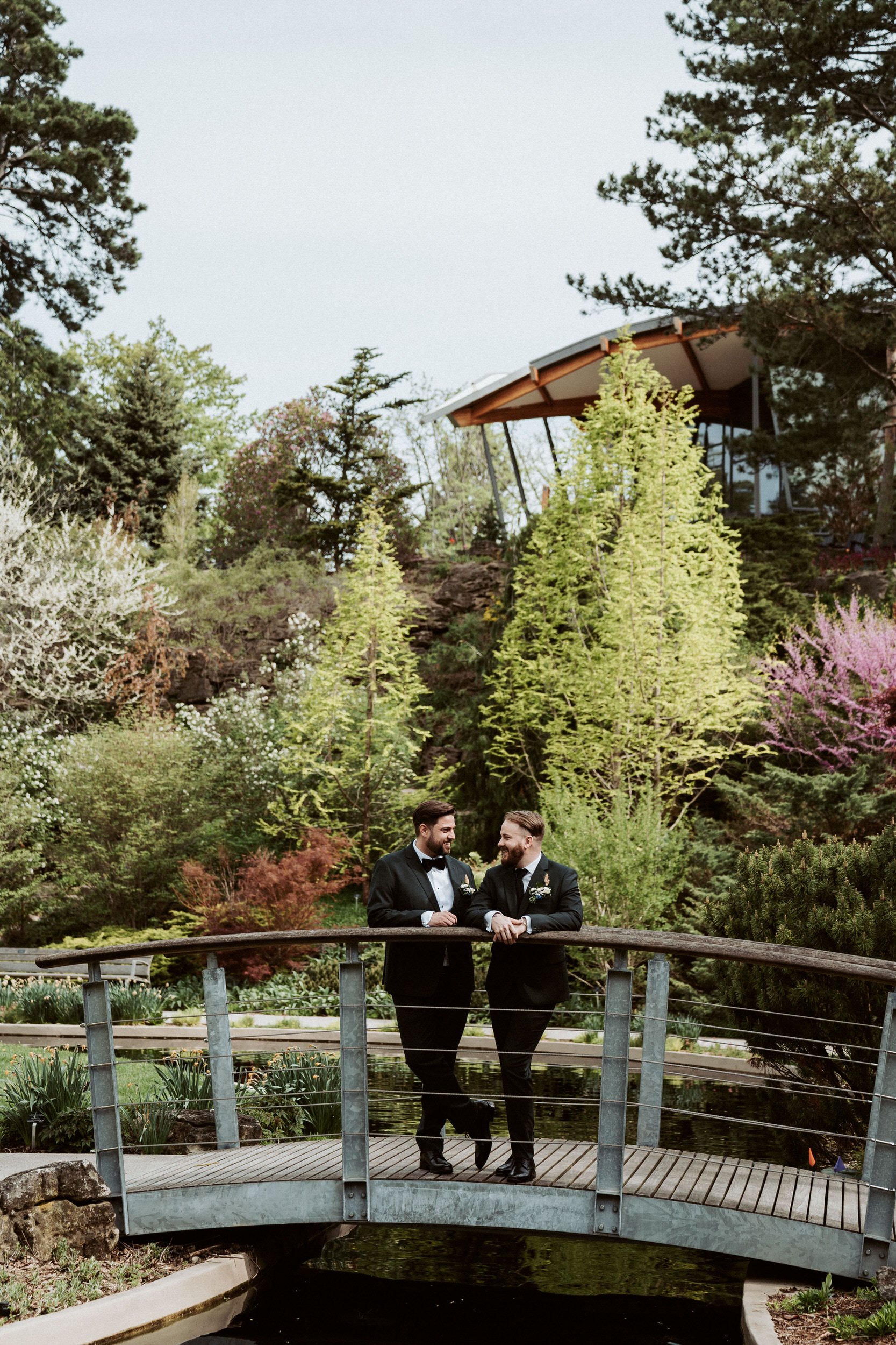 Two men dressed in tuxedos standing on a small bridge in a lush garden, smiling at each other with trees and a modern house in the background.