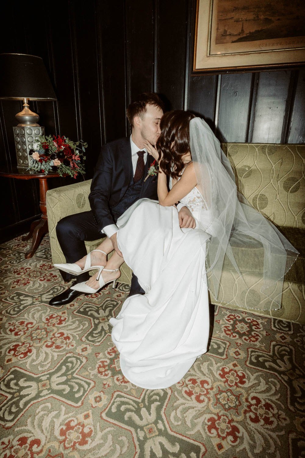 A bride and groom sitting on a vintage sofa, sharing a kiss, with the bride sitting on the groom's lap, in a warmly lit room with dark wood paneling, a patterned carpet, and a side table with a lamp and floral arrangement.