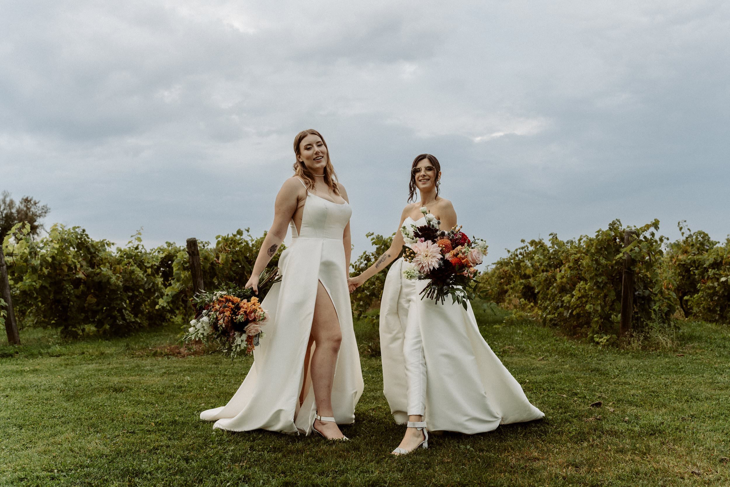 Two women in wedding dresses holding bouquets of flowers, standing hand in hand outdoors in a grassy area with vineyard rows, under a cloudy sky.