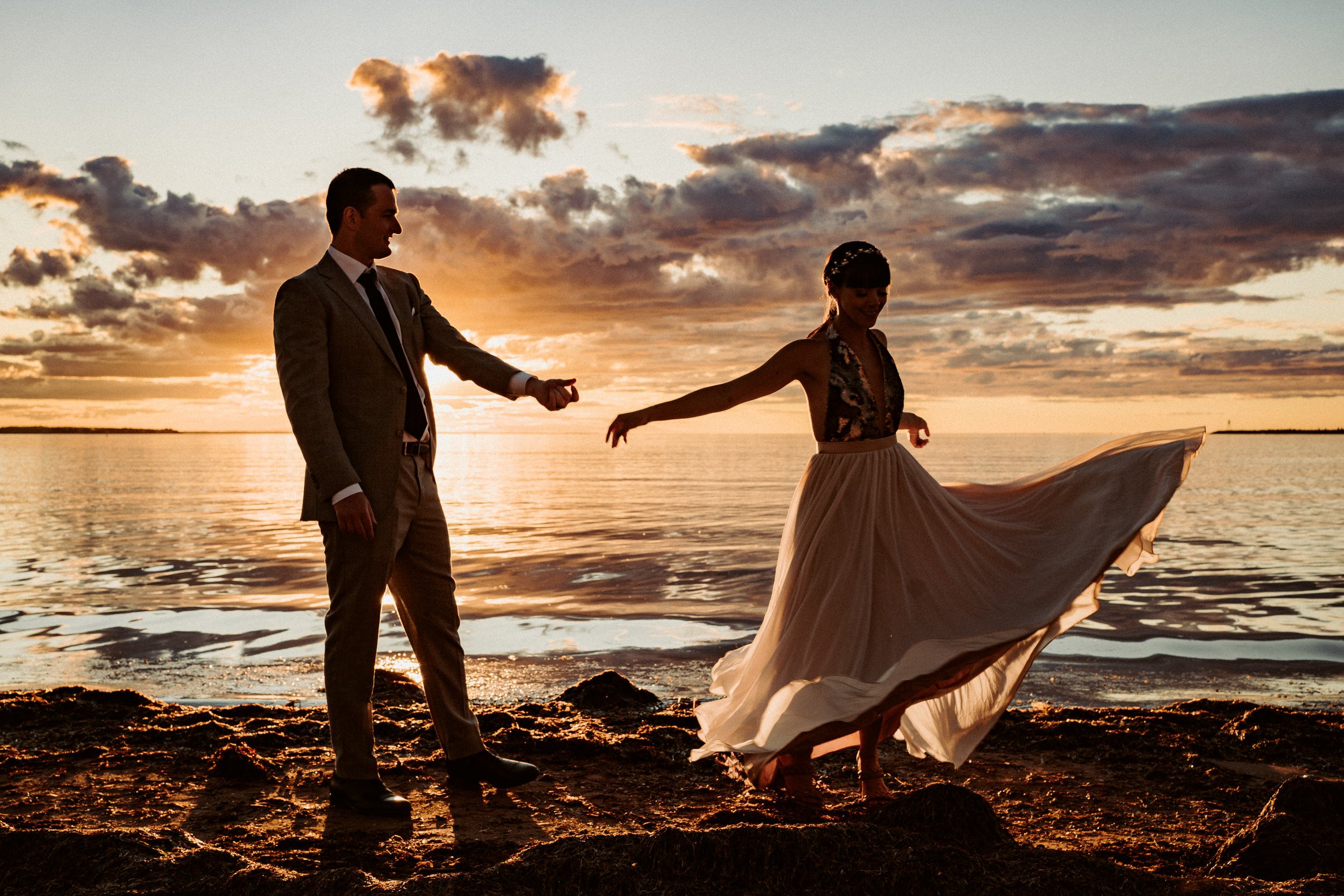 A man and woman stand on the beach at sunset, holding hands and dancing, with the woman's flowing dress billowing in the wind.