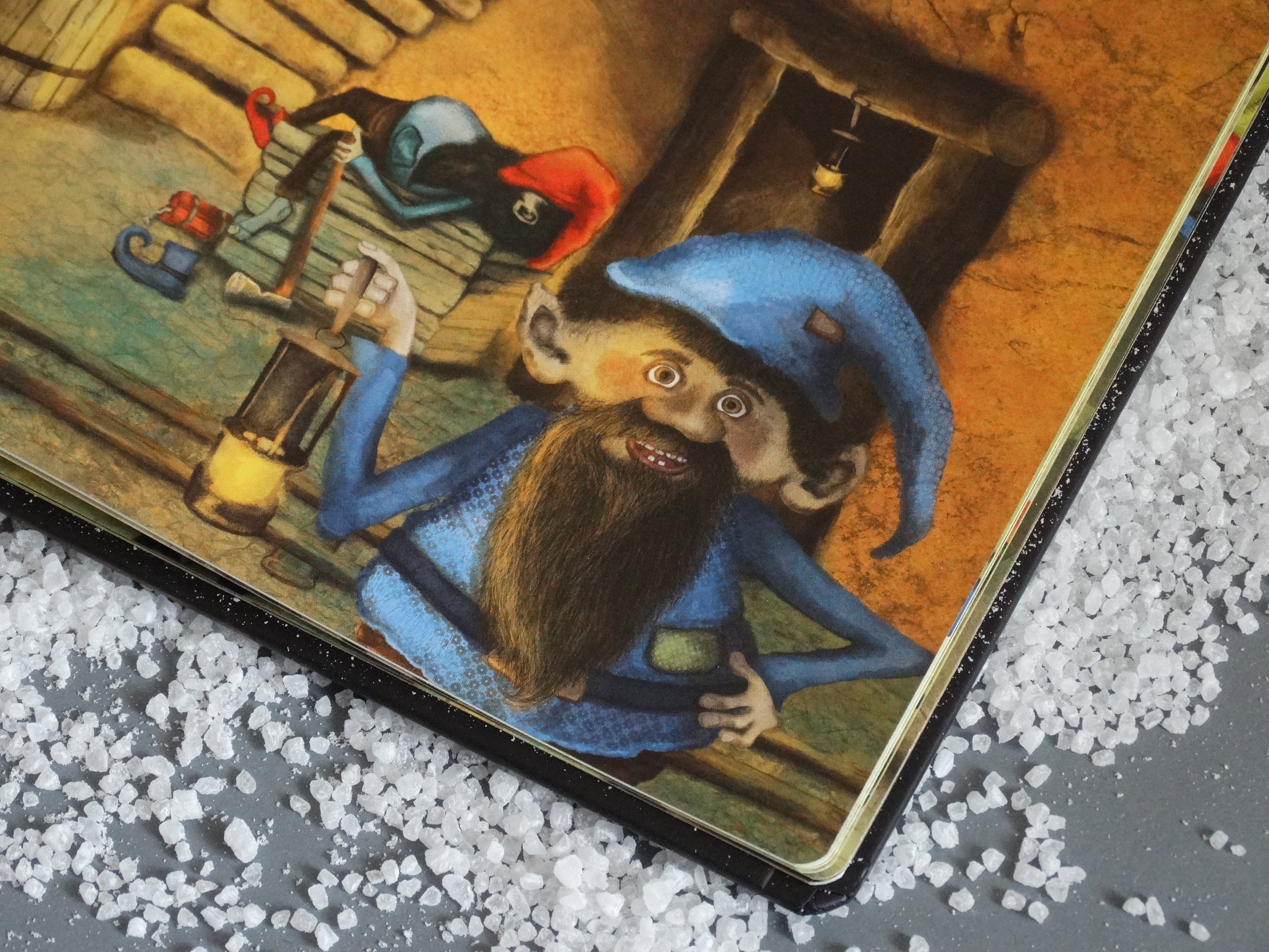 Illustration of a dwarf wearing a blue outfit and hat, holding a lantern, with a bearded face, set in a cave-like environment. Nearby are clothing items and salt crystals are scattered around.