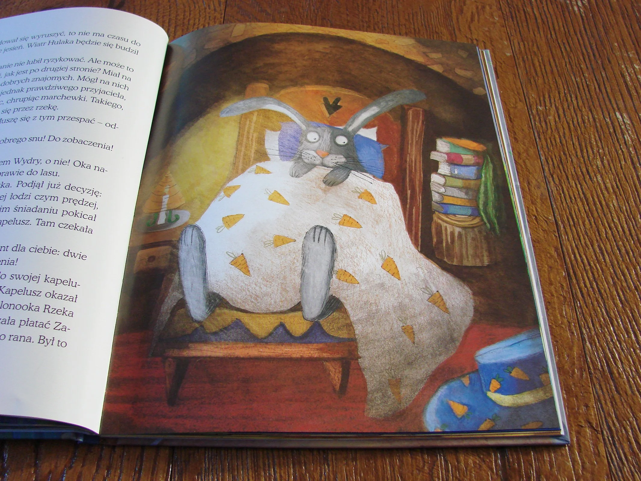 Illustrated page of a book showing a rabbit under a carrot-patterned blanket inside a cozy room, with a bedside table and books.