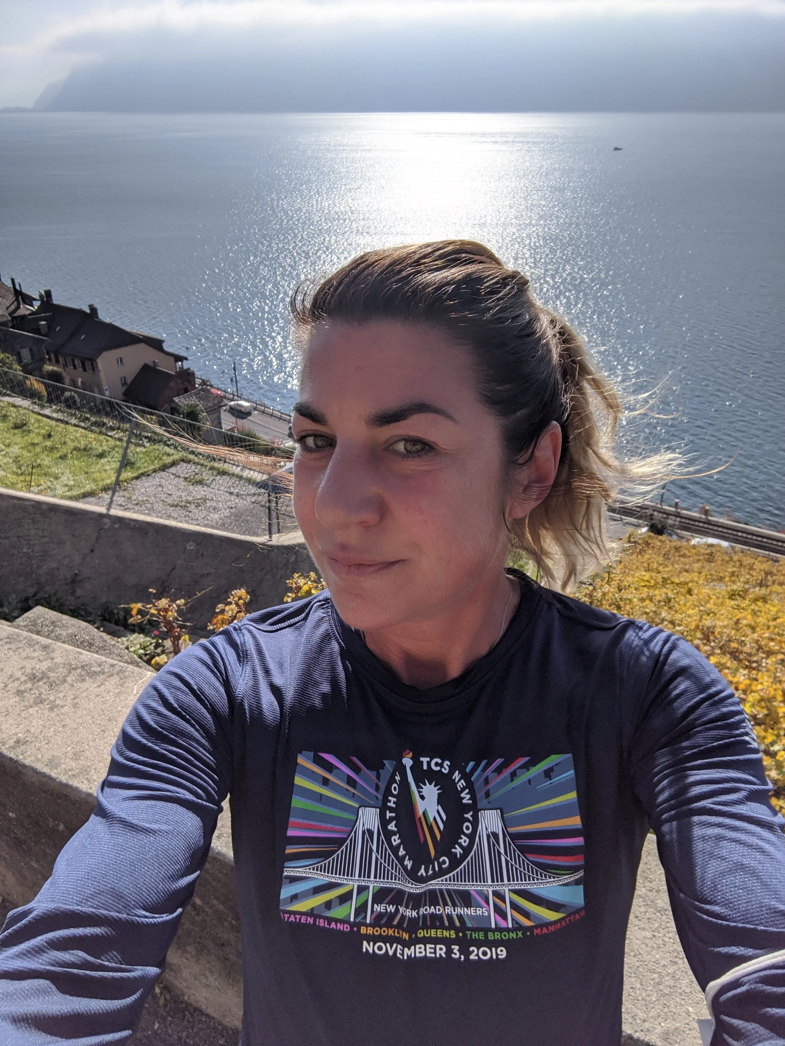 A woman taking a selfie outdoors on a sunny day with a large body of water and a boat in the distance. She is wearing a black race shirt with colorful text and graphics.