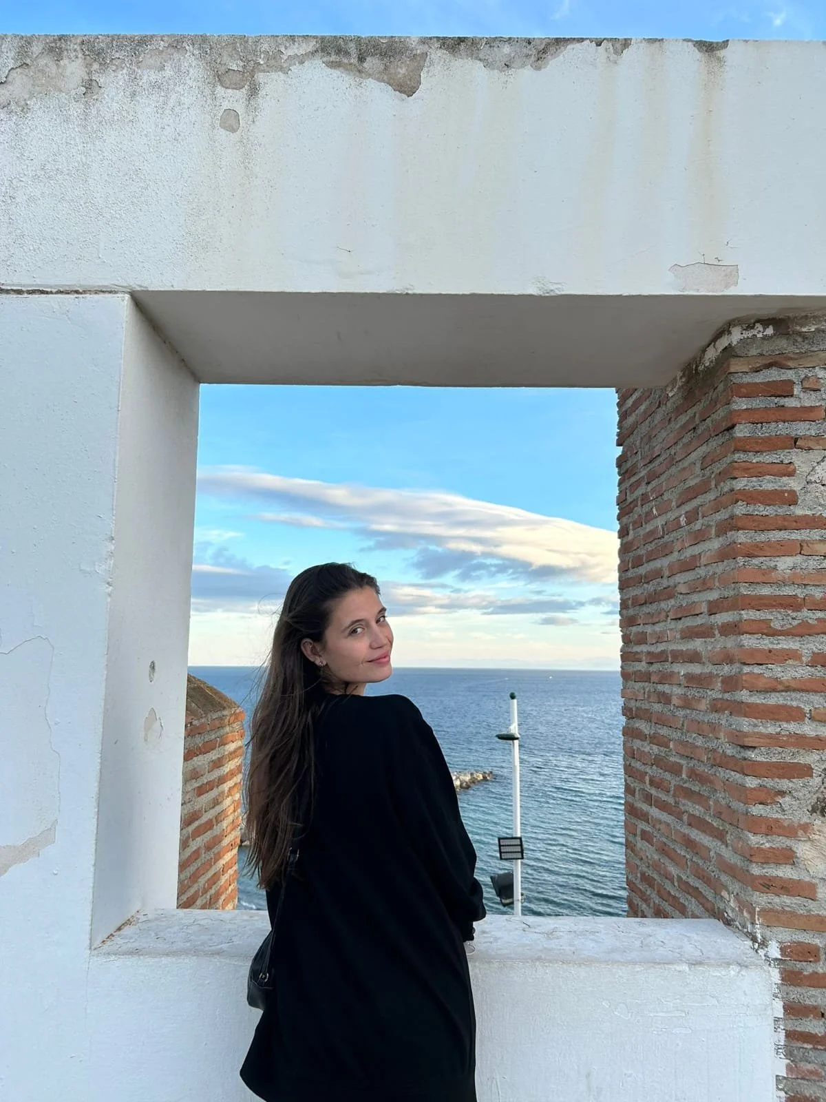 A young woman with long brown hair standing next to a white and brick wall frame at the beach, with the ocean and a cloudy sky in the background.