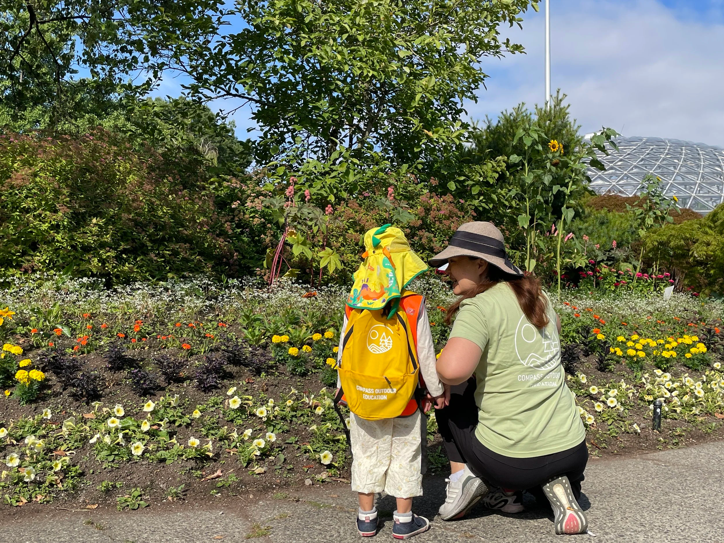 A woman kneeling beside a young girl in front of a flower bed with a large, modern geodesic dome in the background.