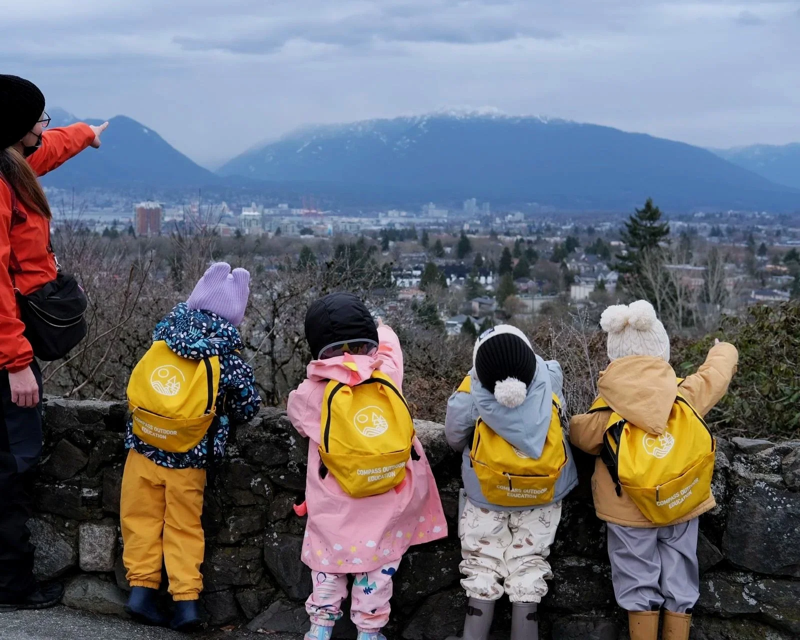 Group of children and an adult looking at a mountain view, pointing towards the mountains, with a cityscape in the background, during cloudy weather.