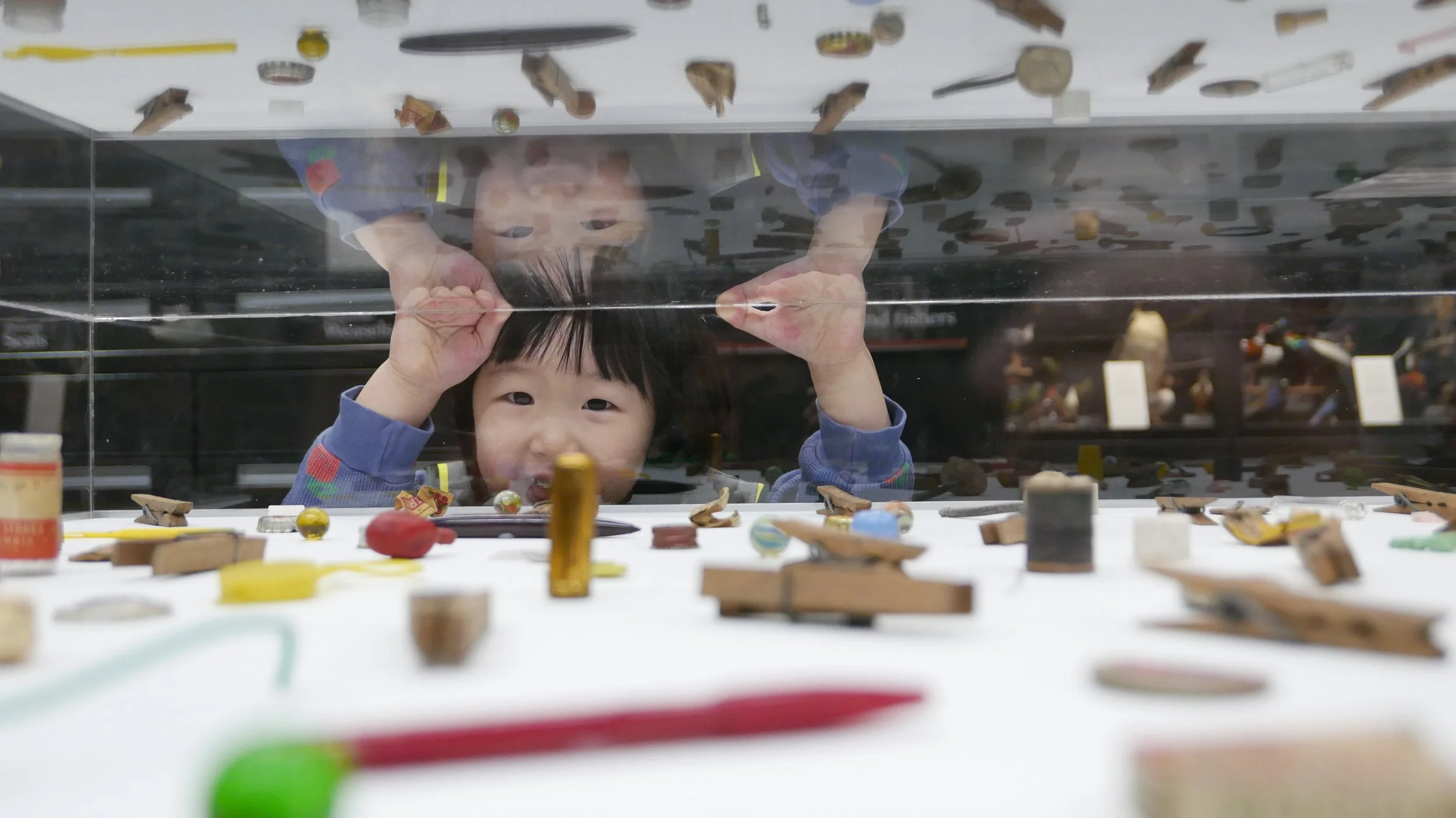 A young Asian child with black hair looking through a glass display case of small, colorful artifacts and toys, smiling and reaching with both hands.