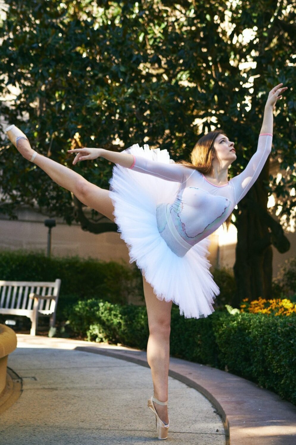White ballerina with long brown hair poses in arabesque on pointe in a garden wearing a white rehearsal tutu