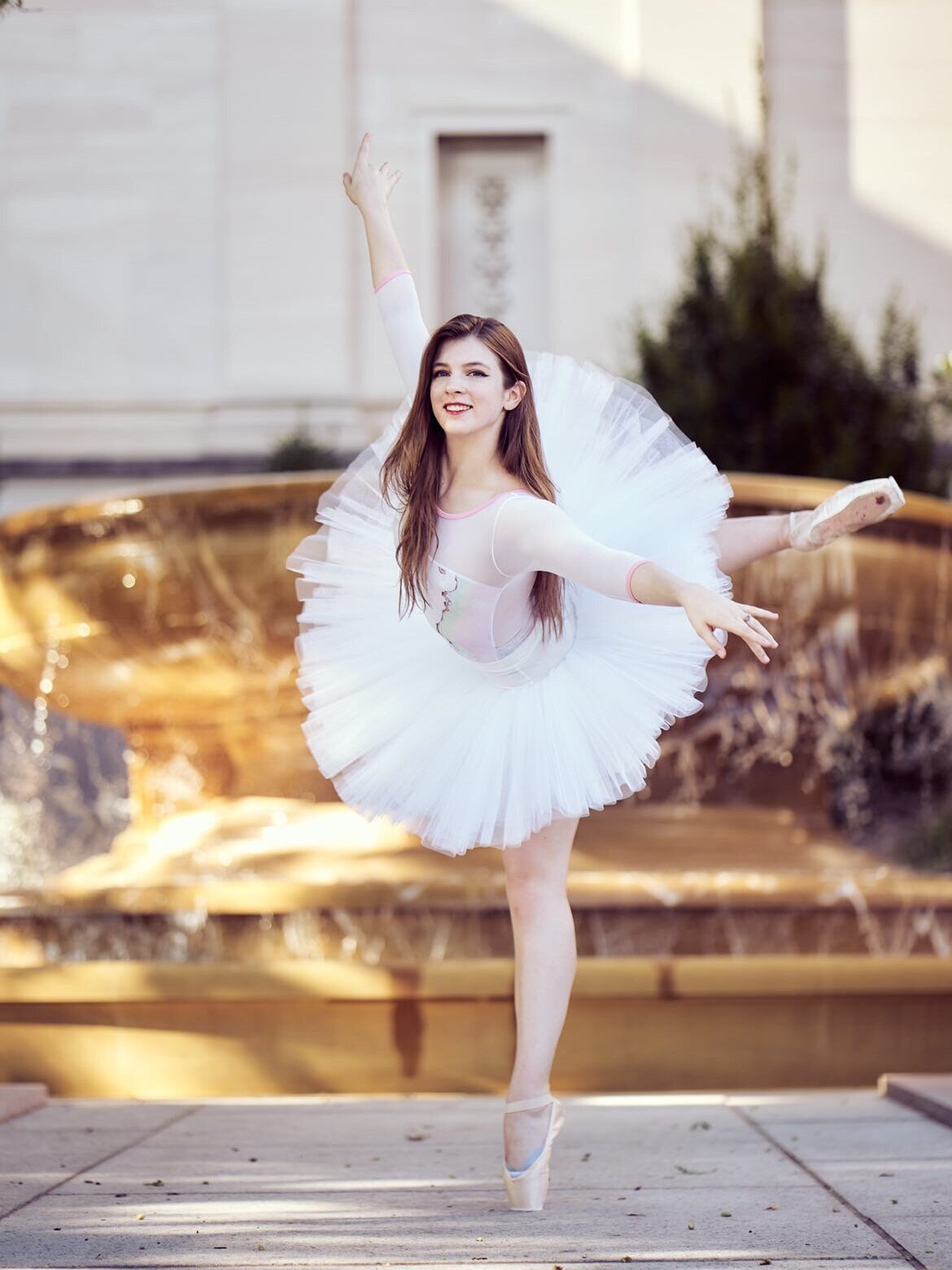 White ballerina with long brown hair poses in an attitude derrier en pointe in a white Bluebird Dancewear Custom Tutu against a yellow brown background