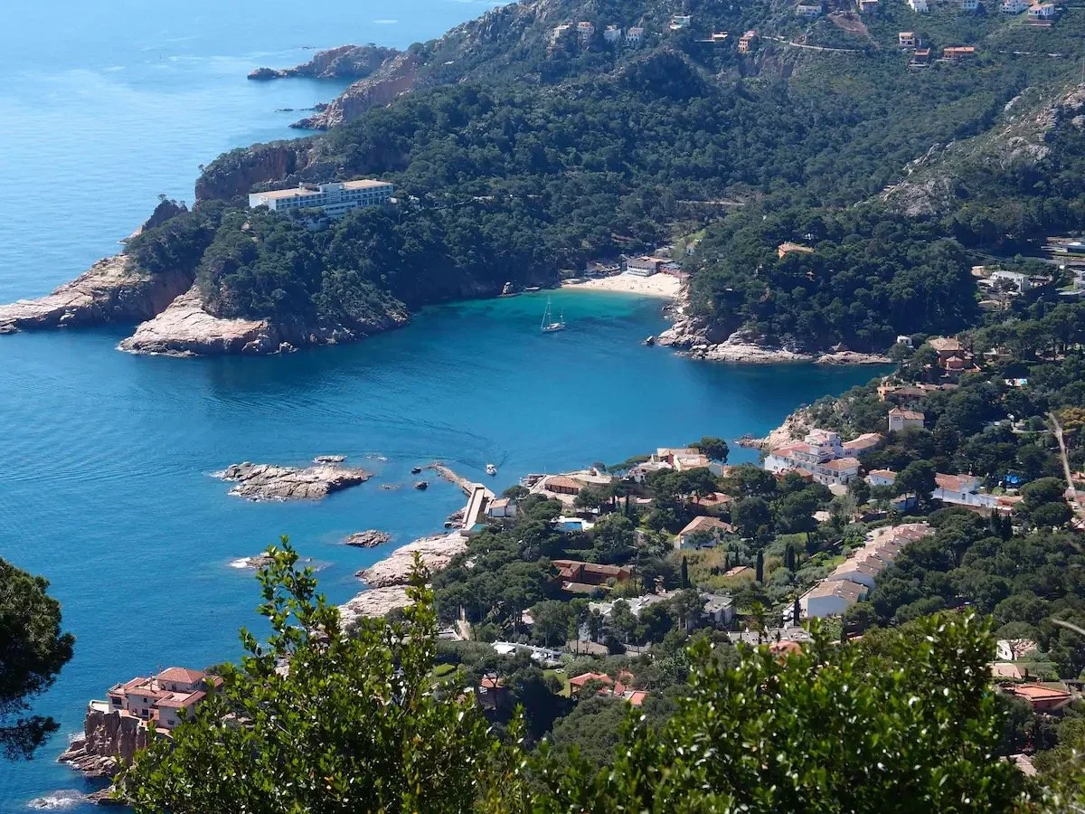 Vue aérienne d'une petite baie entourée de collines verdoyantes, avec des maisons et un voilier sur l'eau bleue.