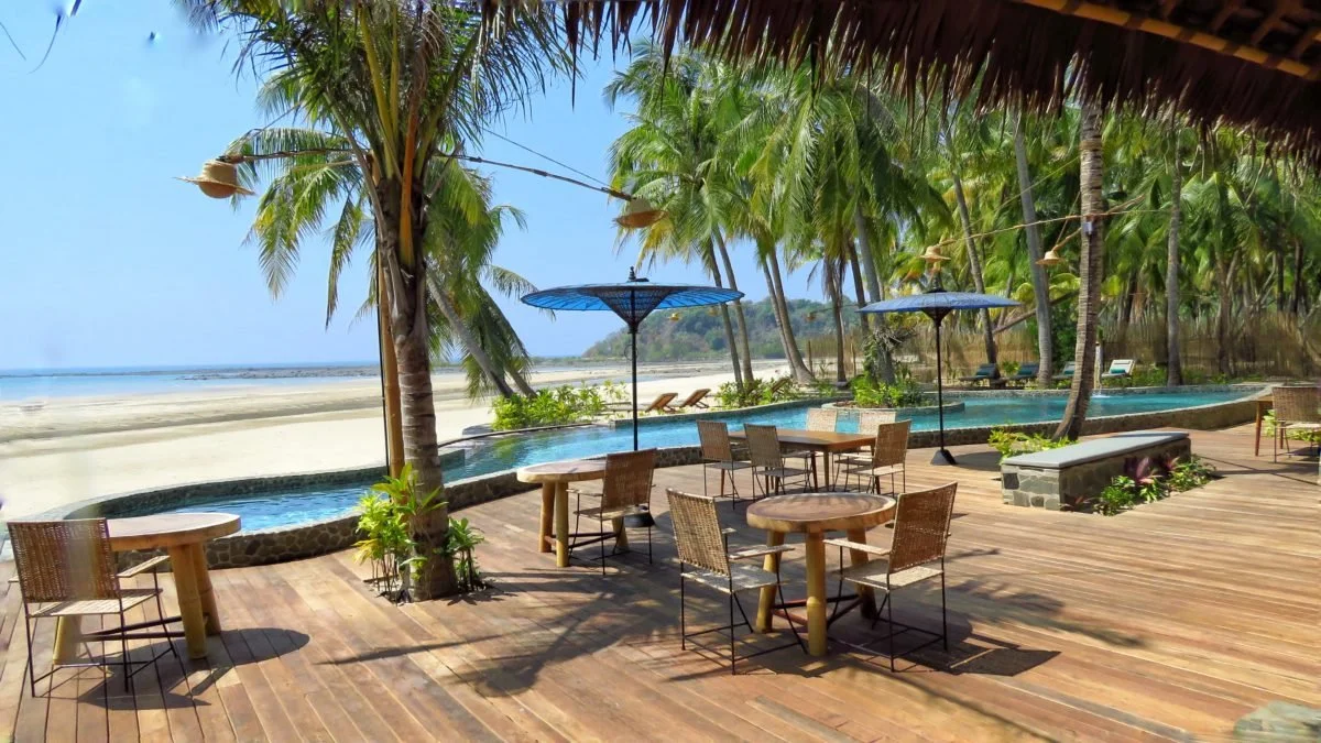 Terrasse en bois avec des tables rondes, des chaises en osier, des parasols bleus, palmiers et piscine en fond, plage de sable et mer.