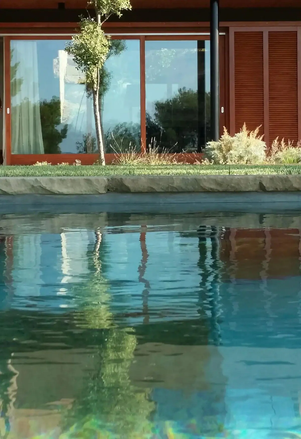 Une maison moderne avec de grandes fenêtres en verre, entourée d'arbres, de buissons et d'un étang ou une rivière avec une surface légèrement ondulée.