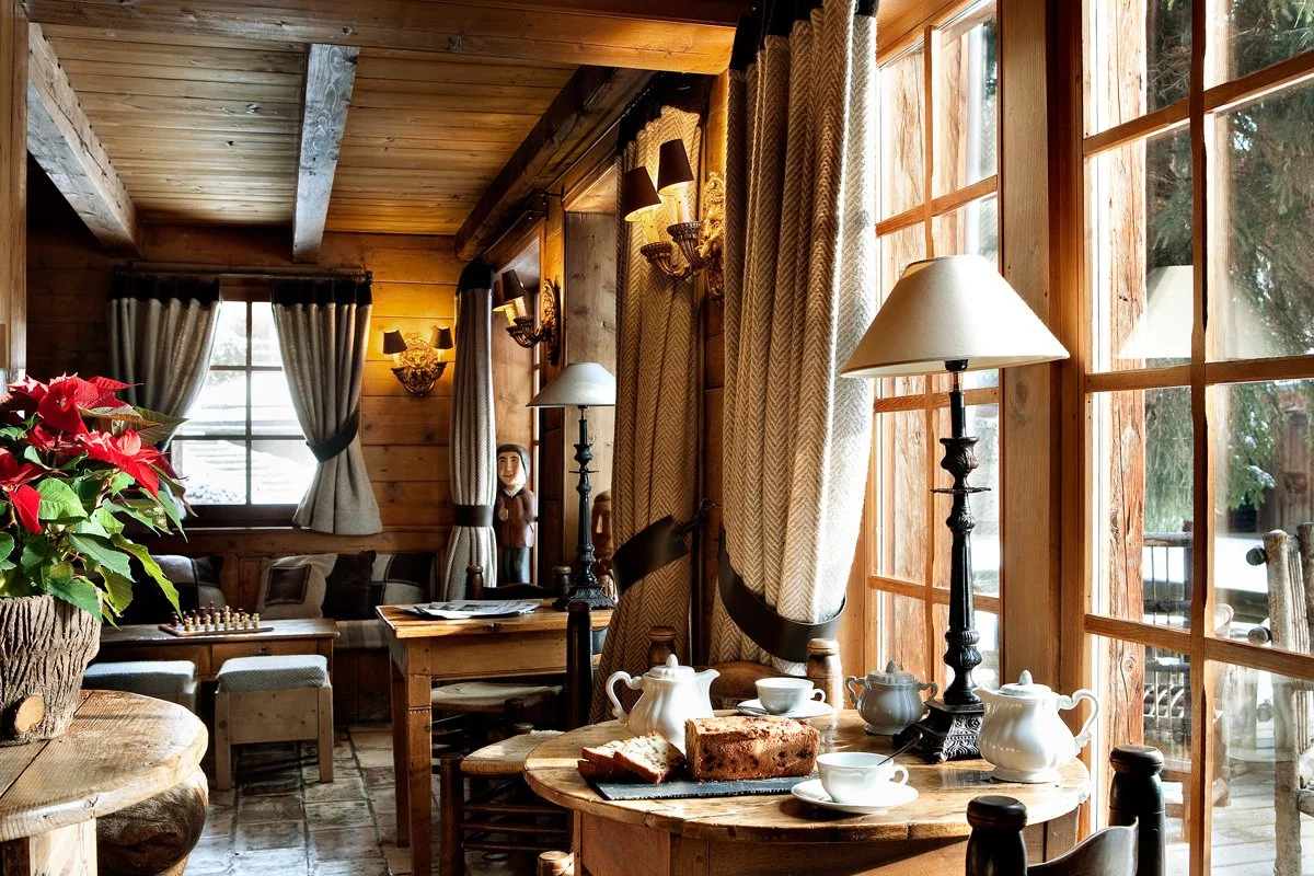 Intérieur d'une cabane en bois avec une table où se trouvent des éléments pour le petit déjeuner, comme du pain, du thé, des tasses, et des lampes, avec de grandes fenêtres en bois montrant l'extérieur.