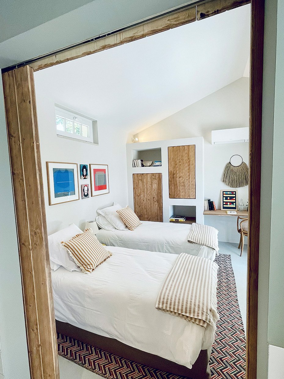 Chambre avec deux lits simples, décoration moderne avec tableaux colorés, bureau avec ordinateur et chaise, étagères en bois, tapis géométrique, mur blanc et lumière naturelle.