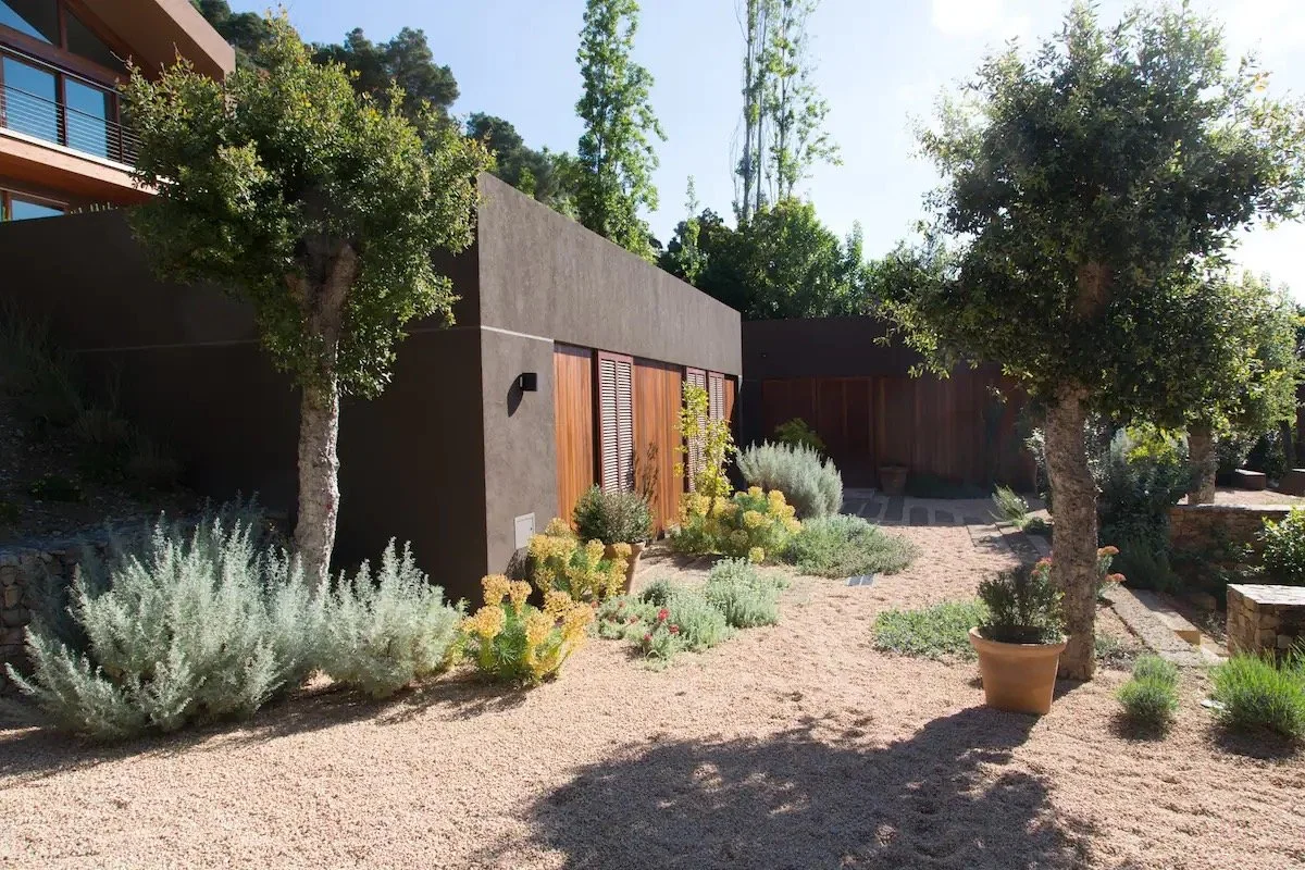Jardin avec arbres, plantes et poteries, bâtiment moderne en arrière-plan, sol en gravier, ambiance ensoleillée.