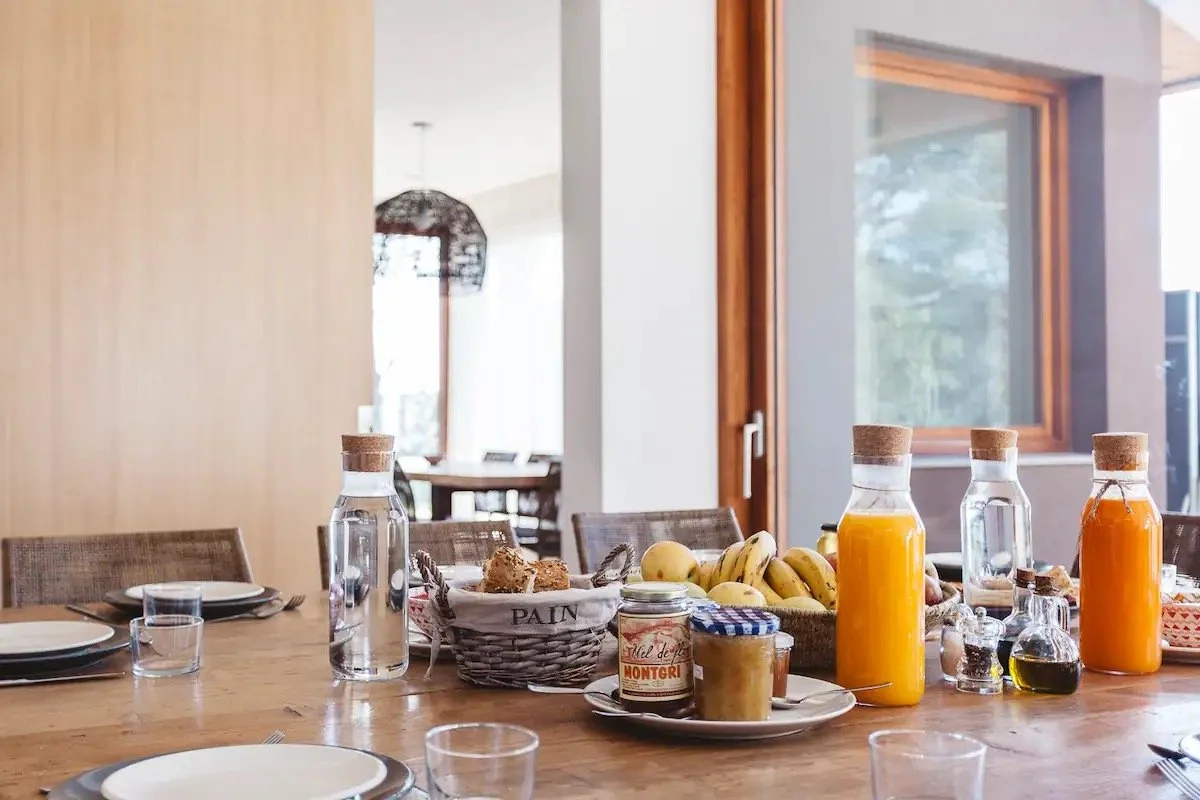 Table de petit-déjeuner avec des bouteilles de jus d'orange, des fruits, du miel, des tartines, et des éléments pour un repas en famille ou entre amis dans une salle lumineuse
