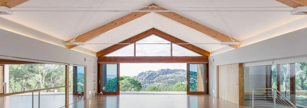 Intérieur d'une salle spacieuse avec grandes fenêtres offrant une vue sur la nature et les montagnes, plafond en bois avec poutres apparentes.