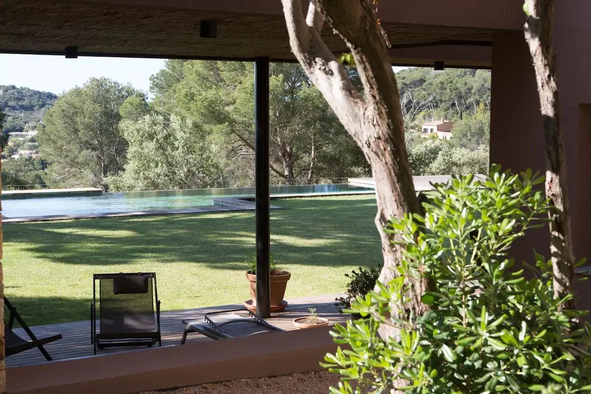 Une vue depuis un espace intérieur donnant sur un jardin avec une pelouse, des arbres et une piscine en arrière-plan, avec des chaises de plage sur le patio.