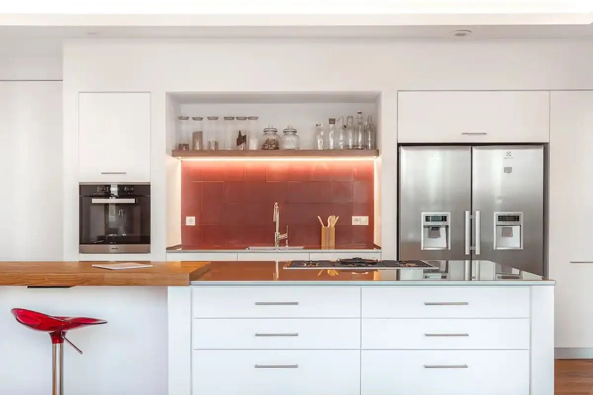 Cuisine moderne avec armoires blanches, réfrigérateur double porte en acier inoxydable, four intégré, îlot avec plaques de cuisson et un rangement en bois, un lavabo avec robinet, étagère ouverte avec bocaux et bouteilles en verre, éclairage sous les