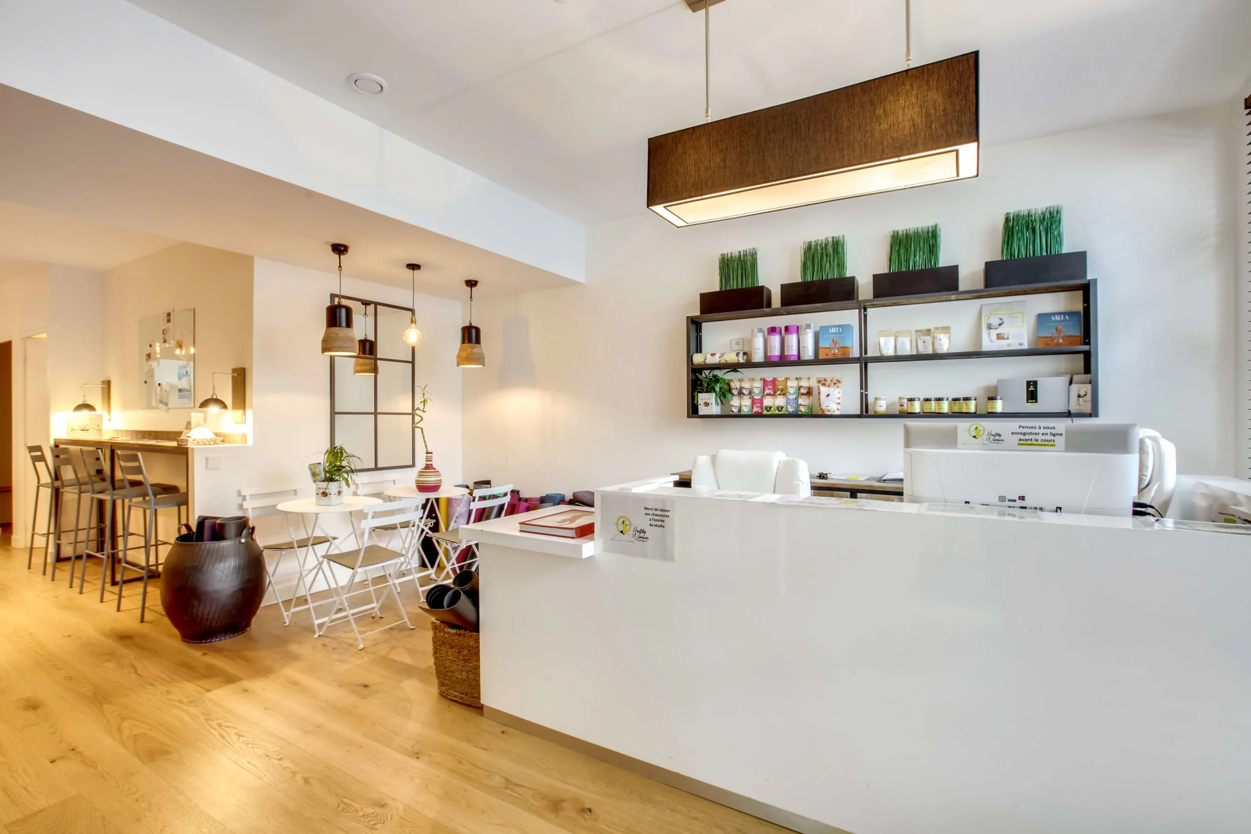 Un intérieur moderne et lumineux, comprenant un comptoir blanc, des étagères avec des produits, des tables et chaises blanches, et des suspensions lumineuses. Le sol est en bois clair et des plantes décorent l'espace.