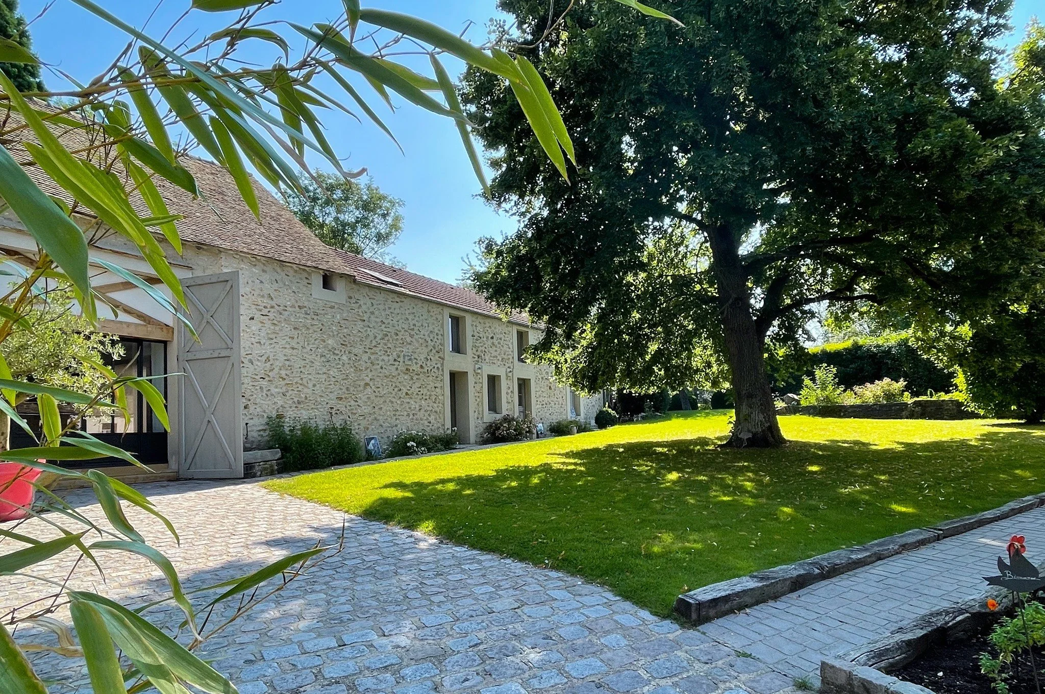 Une maison en pierre avec un toit en tuiles rouges, une porte et plusieurs fenêtres, entourée d'un jardin verdoyant avec un arbre imposant, un chemin en pavés et un ciel bleu en arrière-plan.