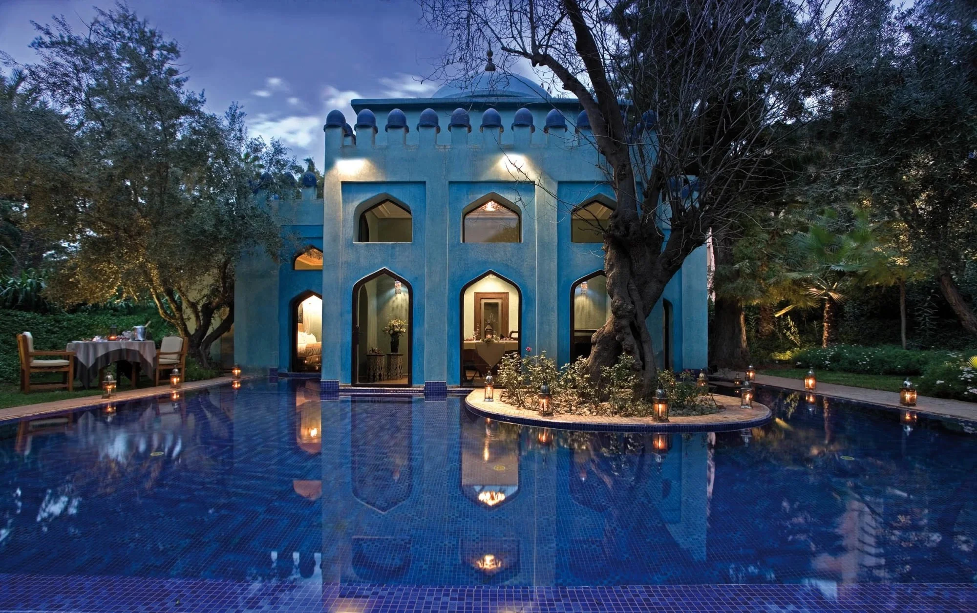 Maison bleue de style architectural marocain entourée de palmiers et arbres, avec une piscine éclairée par des lanternes lors de la soirée.